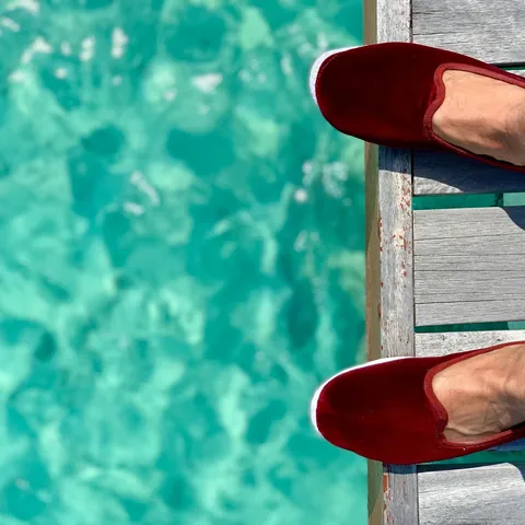 Burgundy velvet slippers on a weathered wooden jetty above turquoise water