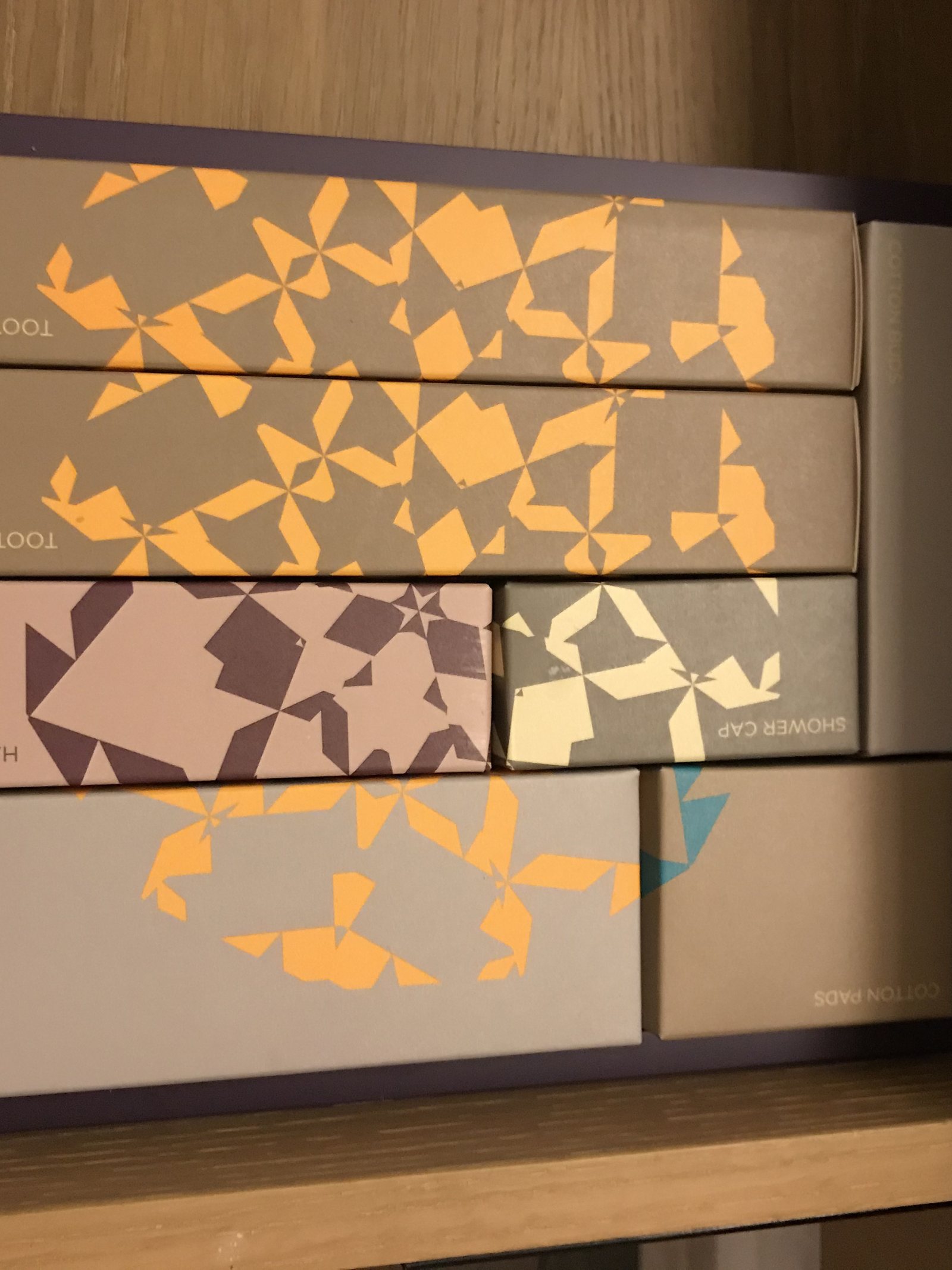 Four amenity boxes in printed card, patterned in orange and purple, on a timber shelf