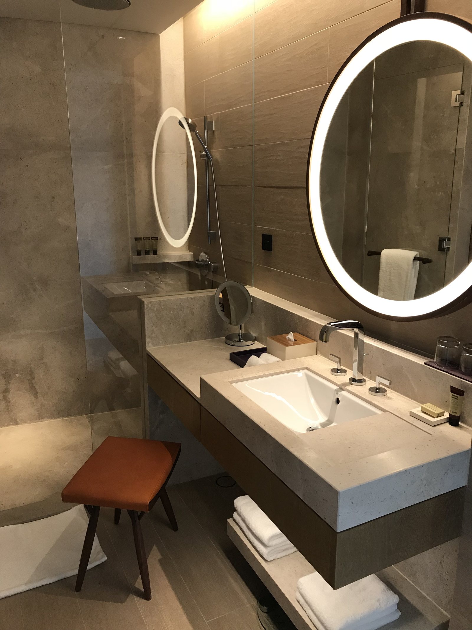 A stone bathroom with a round lit mirror, a long vanity and a stool beside folded towels