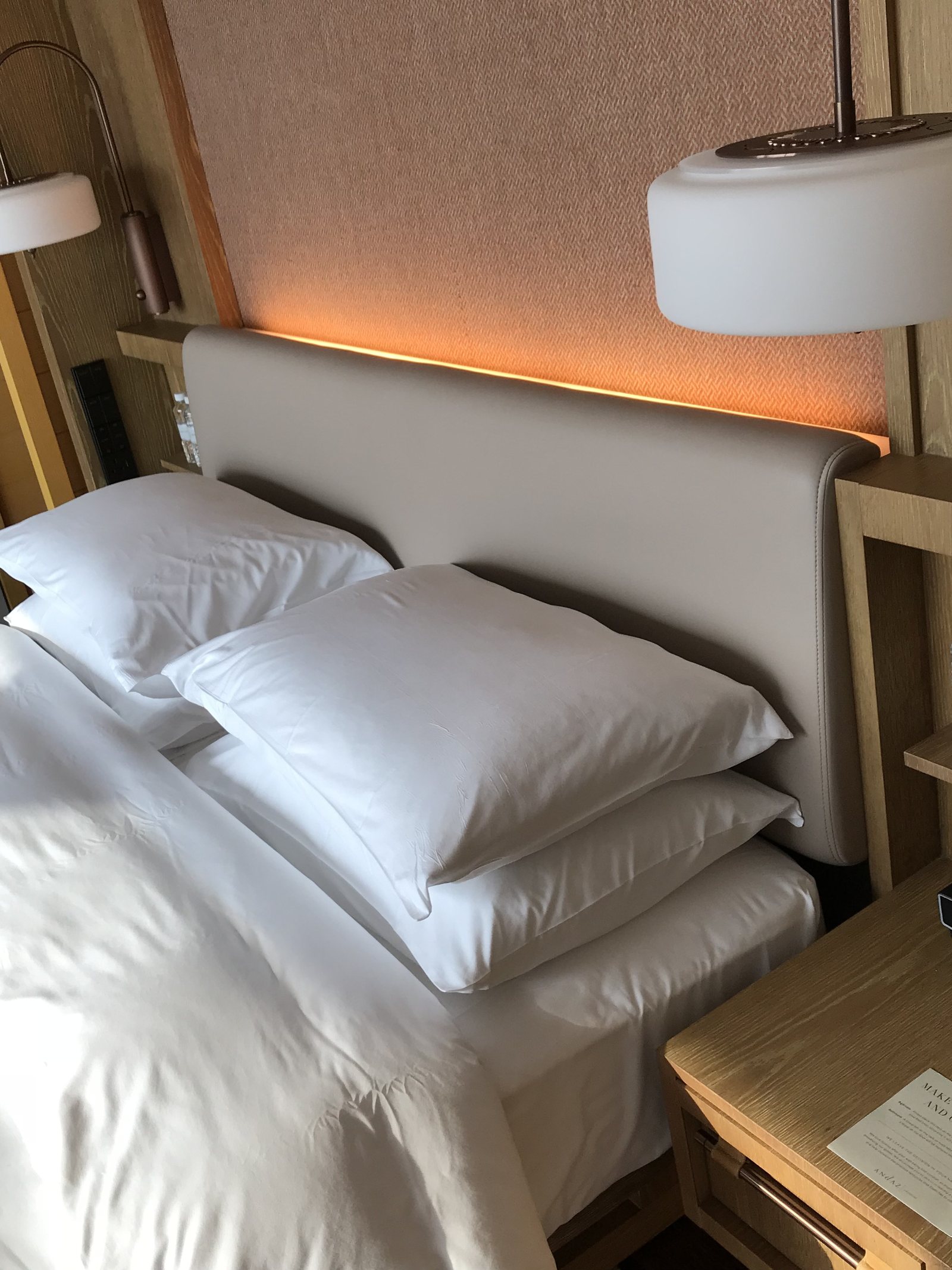 A bed with white linen under a curved headboard, with reading lights and a timber bedside shelf