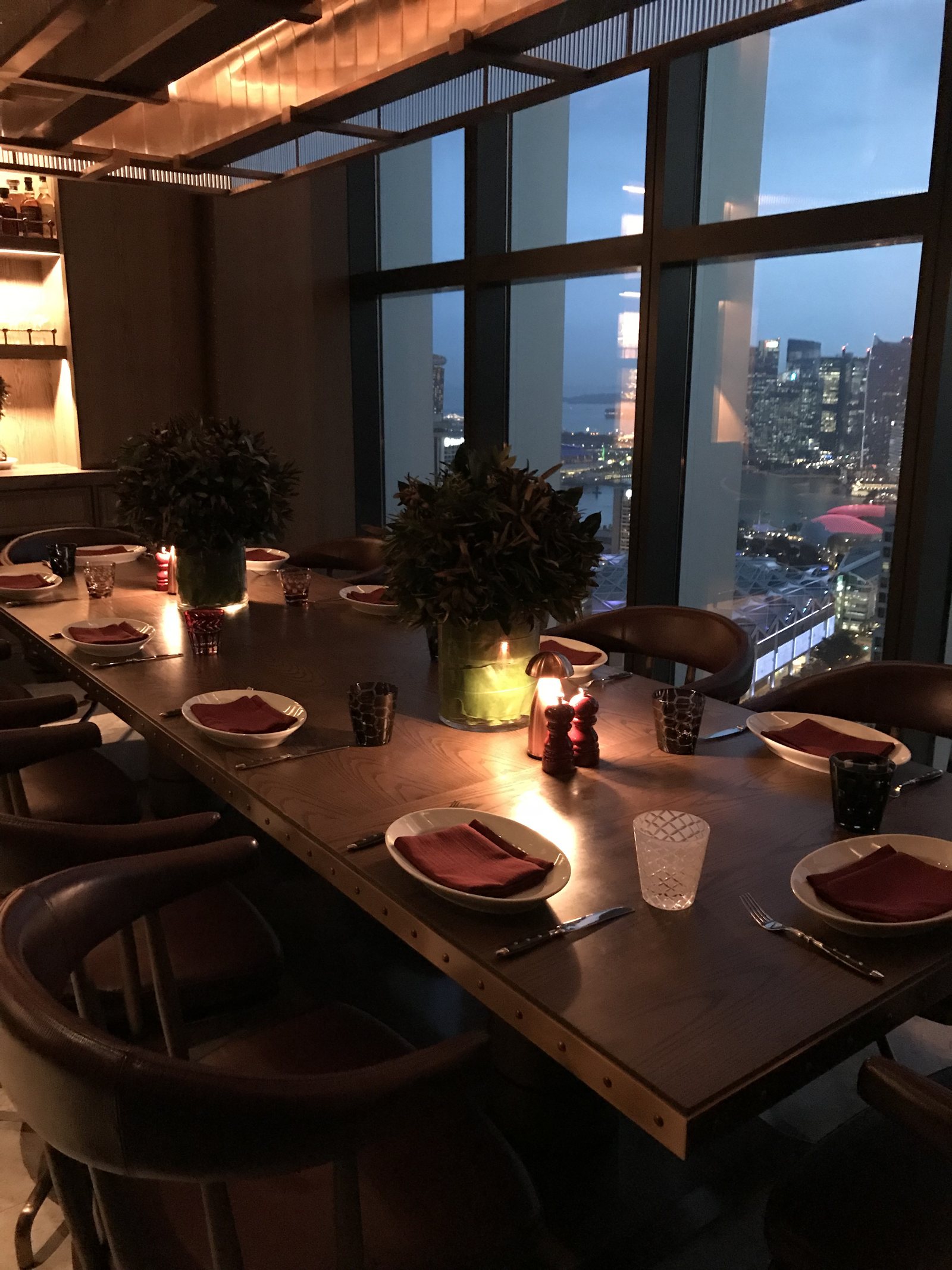 A long timber communal table laid with glasses and candles, the lit skyline behind it