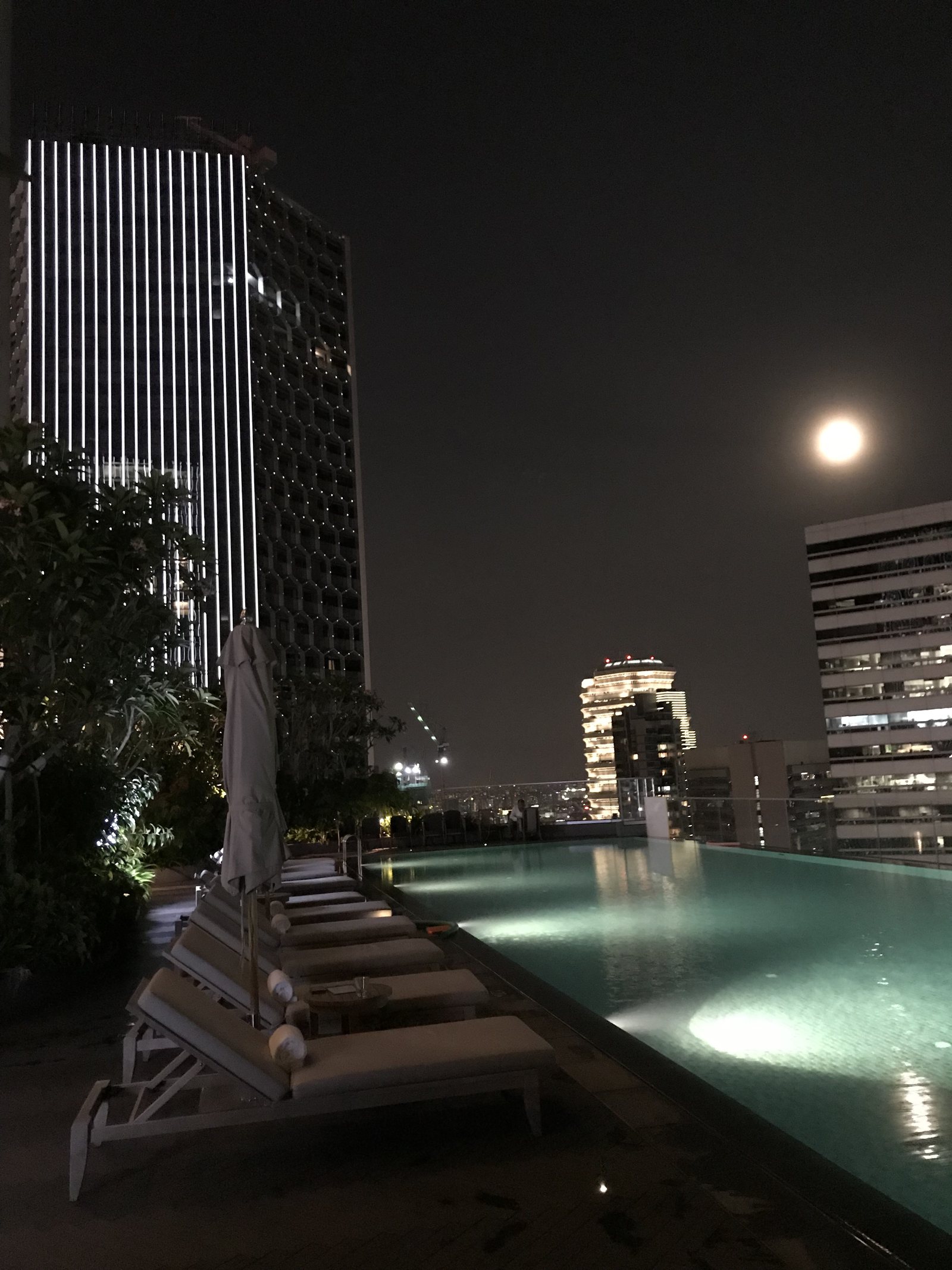 The pool at night with a full moon over the buildings beyond the loungers