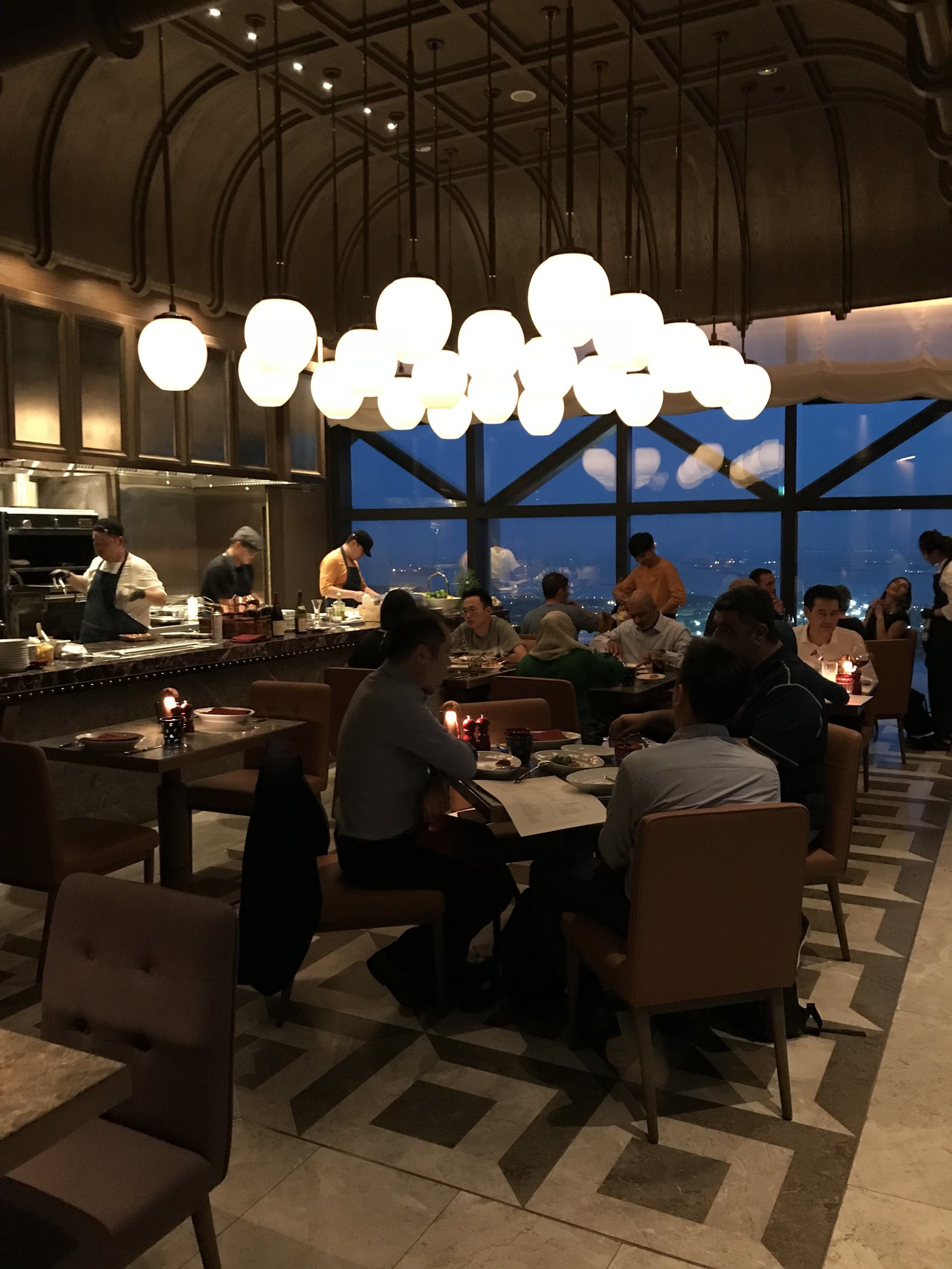 An interior bar with a cluster of globe lights over tables, the city visible through the glass