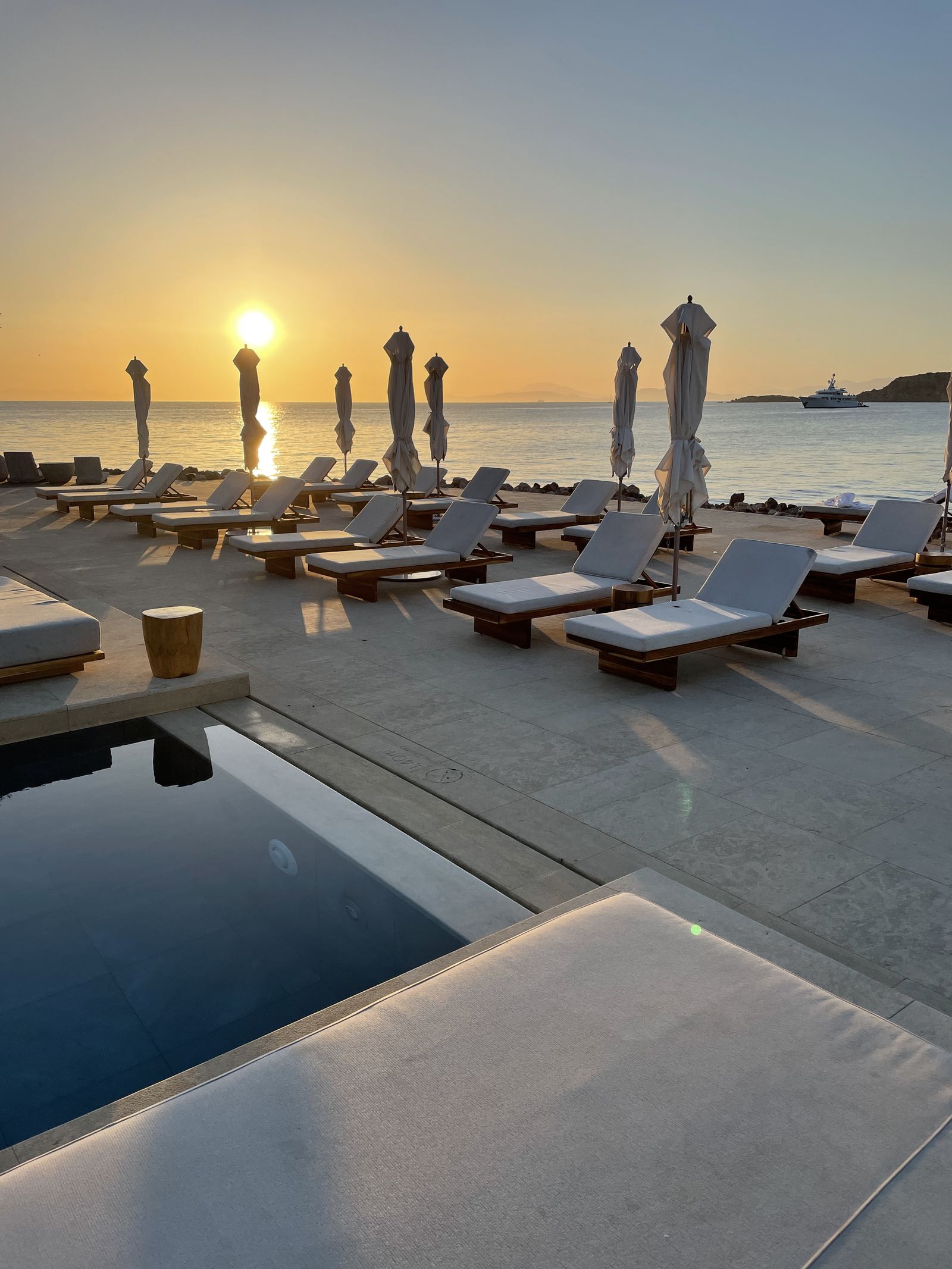 Loungers and folded parasols by the pool with the sun low over the sea