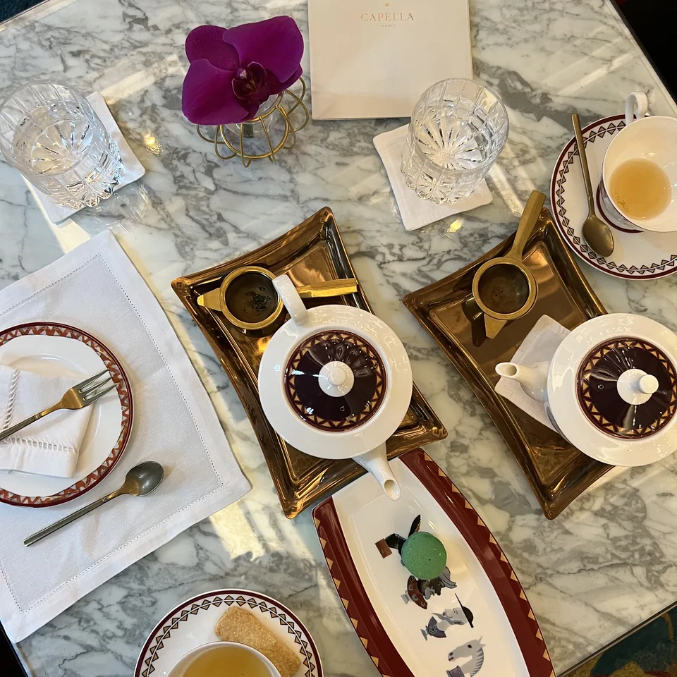 A marble table seen from above, laid with patterned cups, gold trays, a teapot, an orchid and a tray of petits fours
