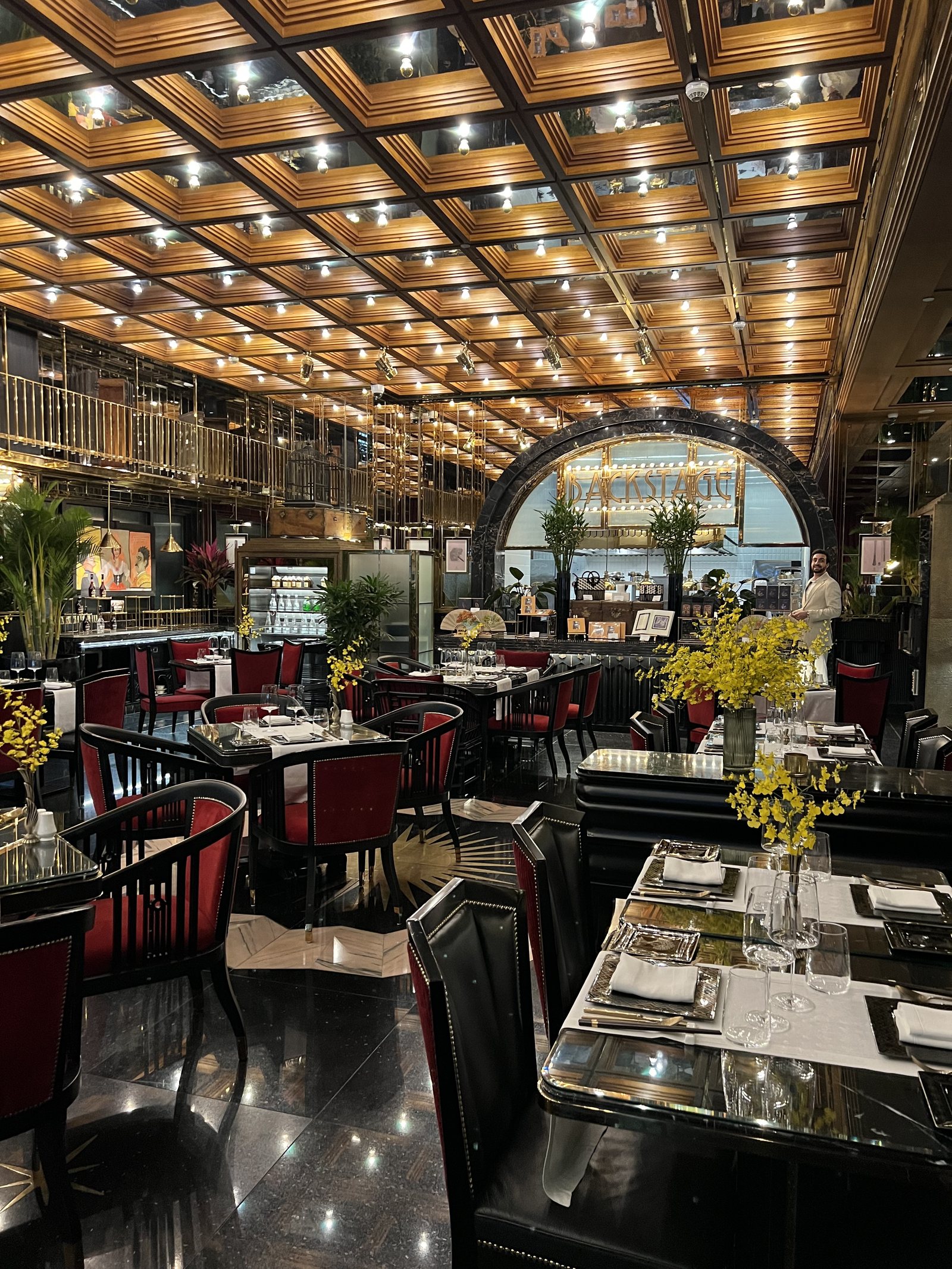 A dining room with red chairs at black tables under a gold coffered ceiling studded with lights
