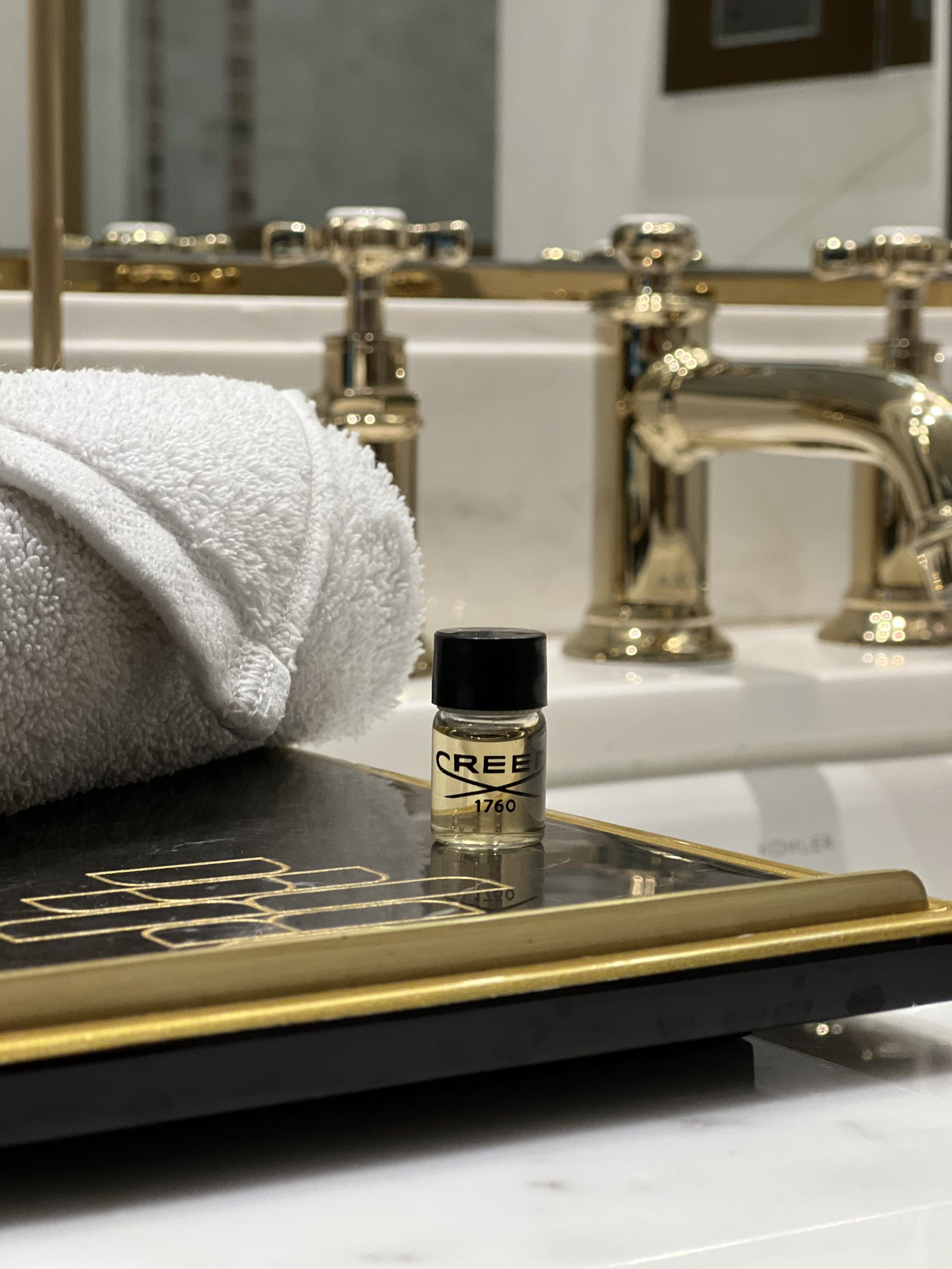 A rolled white towel and a small bottle of Creed on a brass tray across the rim of the bath