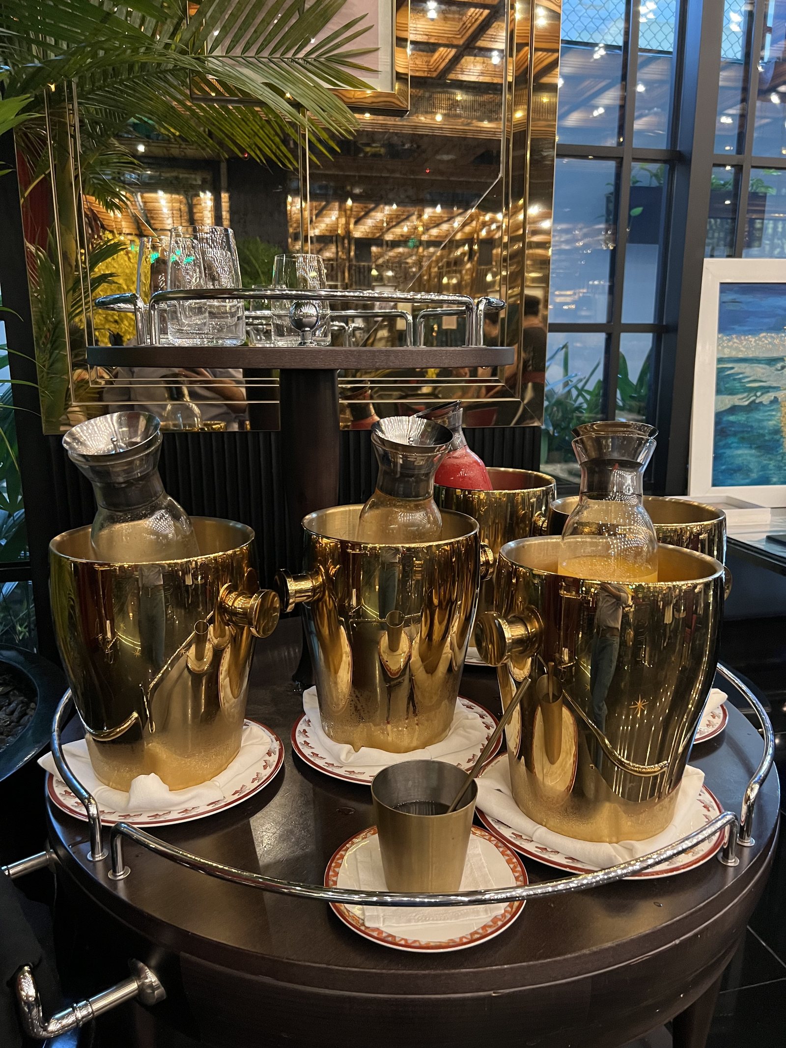 Three brass jugs and a small cup on a trolley set with saucers, in front of a window