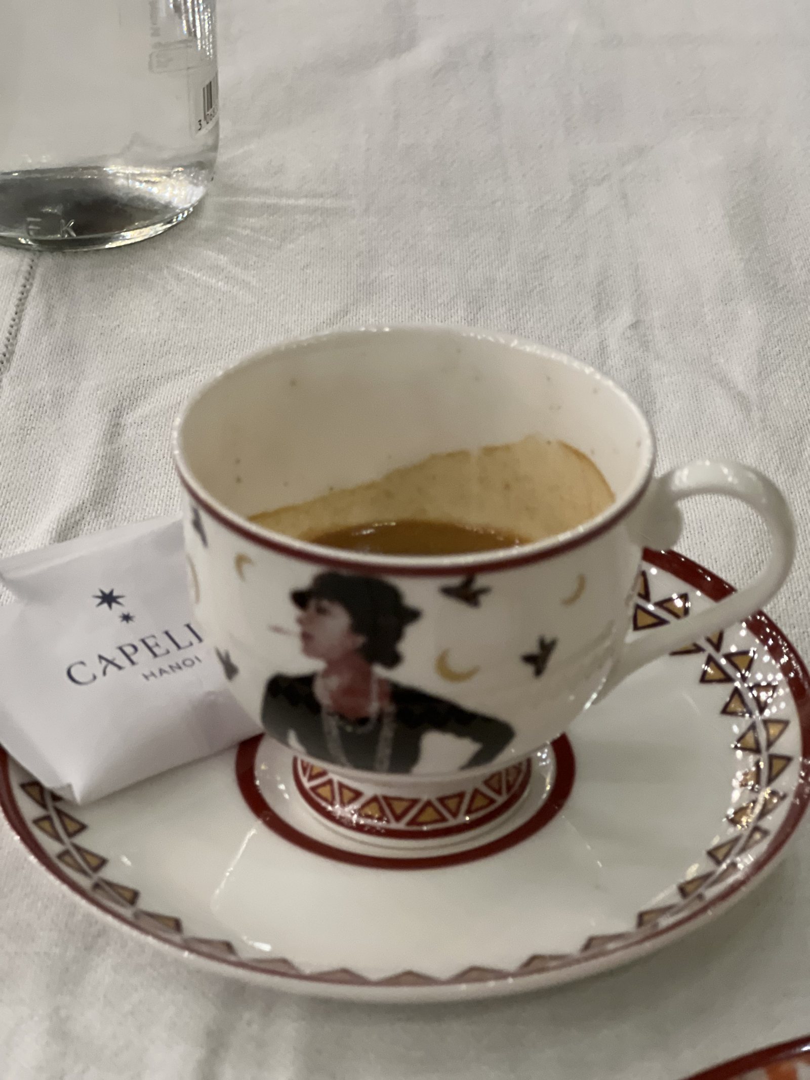 A coffee cup painted with the face of a woman in 1920s dress, on a matching saucer beside a Capella sugar packet