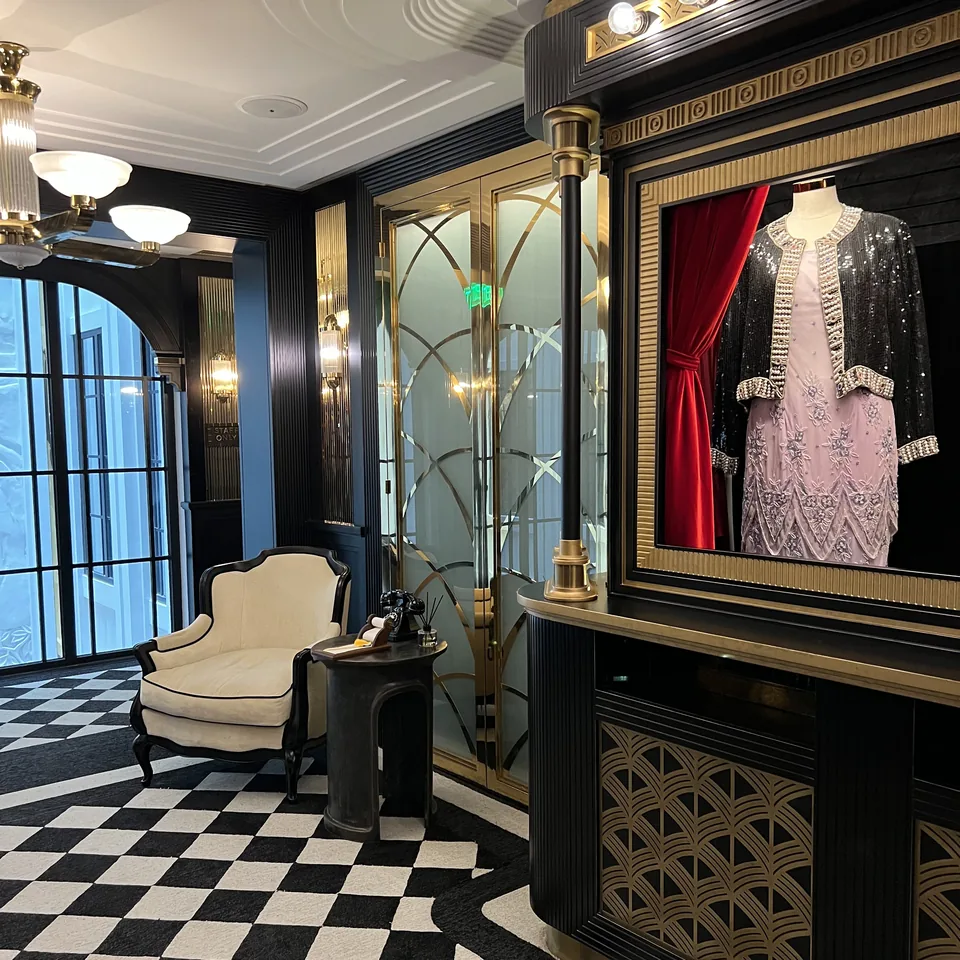 A beaded pink stage costume in a lit glass case beside a red curtain, on a black and white chequered floor
