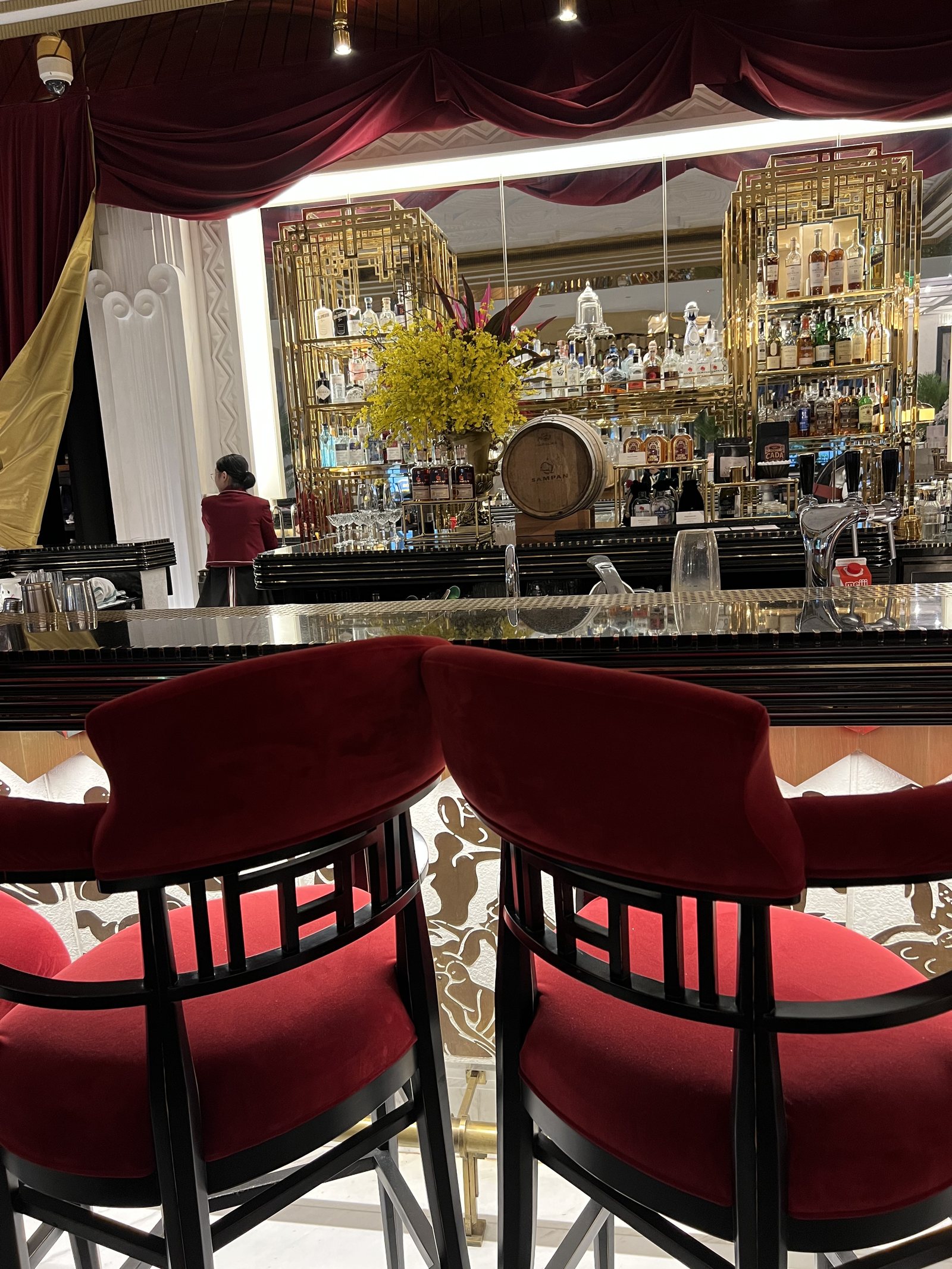 A bar with red velvet stools in front of shelves of bottles under a gilded ceiling