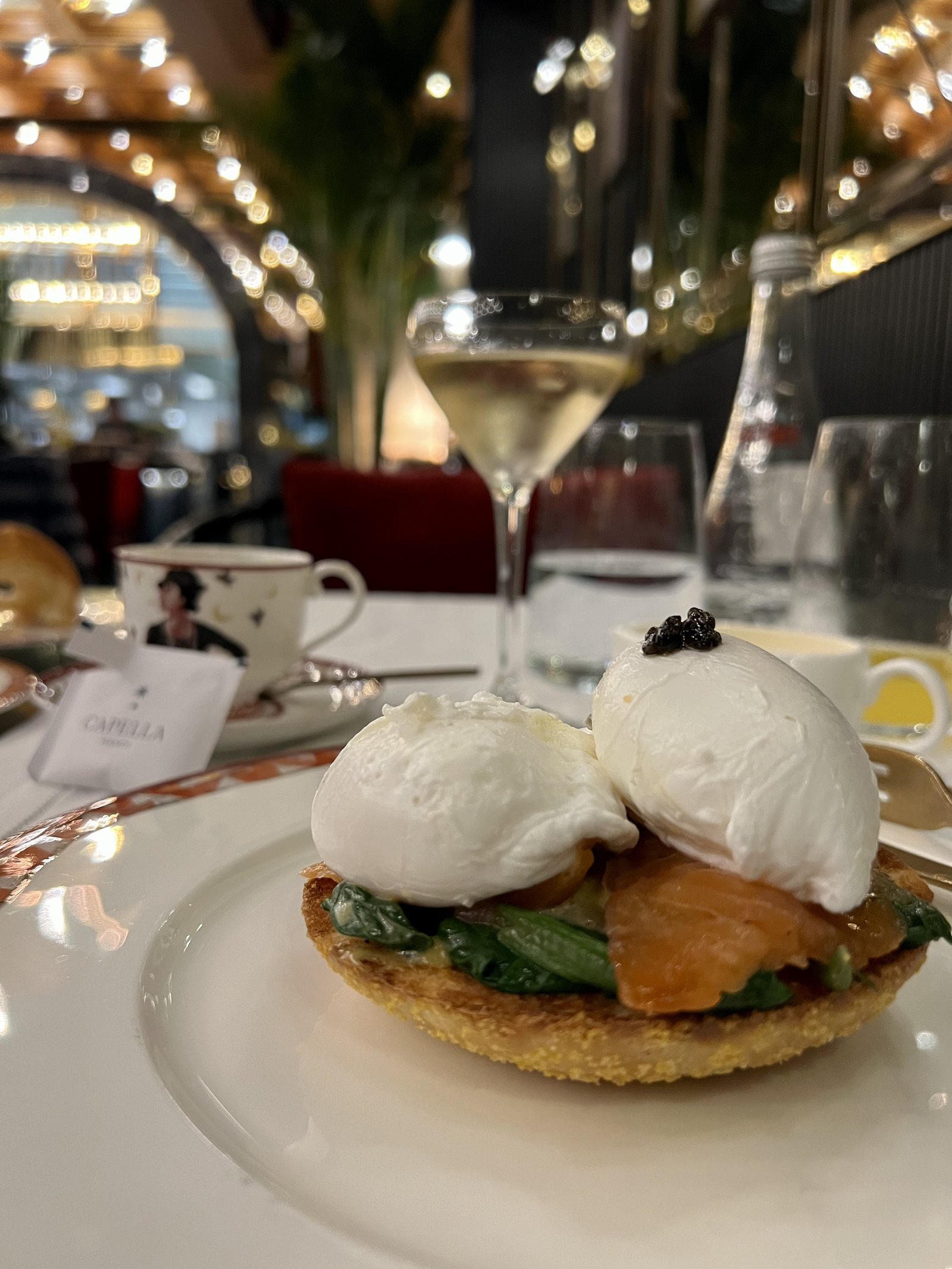 Two poached eggs on salmon and greens on a rosti, on a white plate beside a coupe of champagne