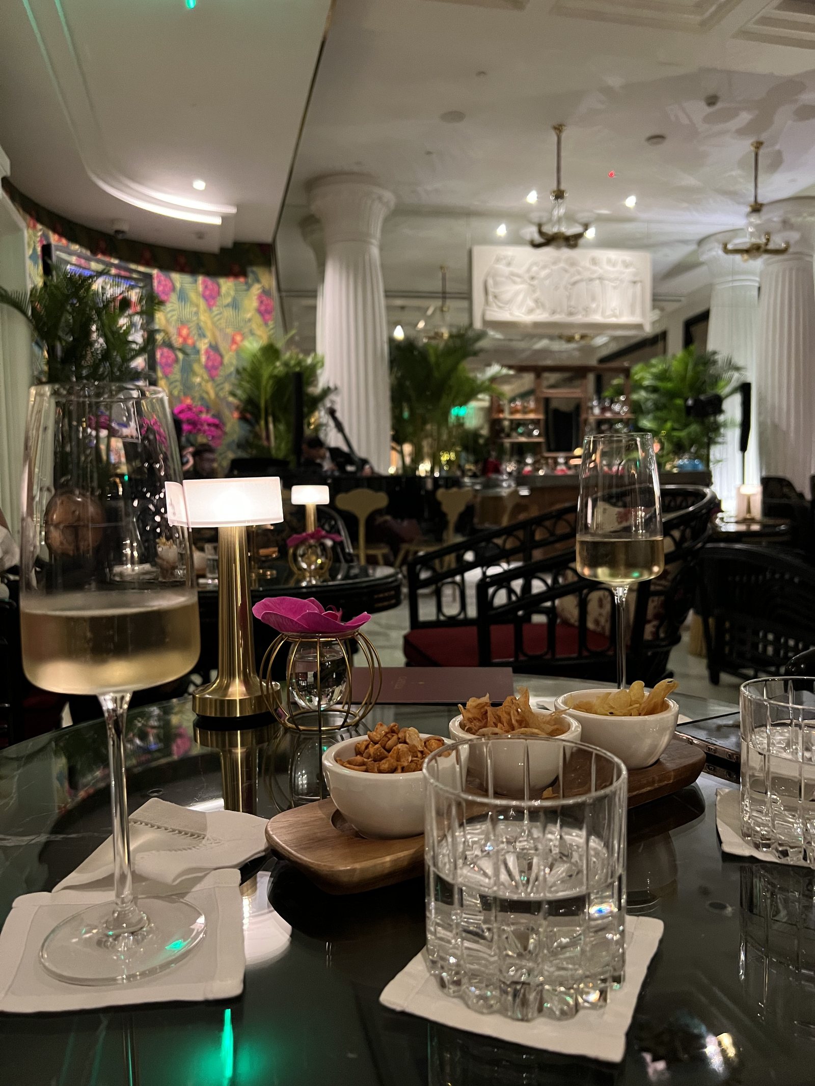 A marble table with two glasses of wine, bowls of nuts and crisps and a water glass, in a lit lounge