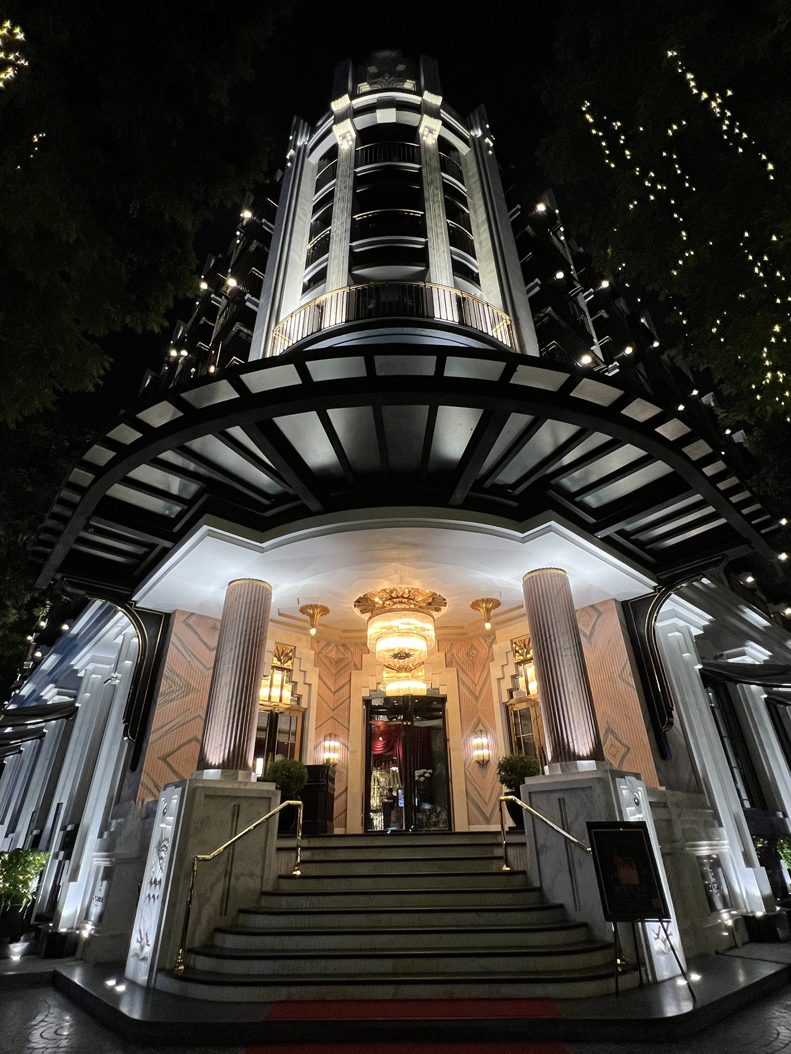 The lit entrance of Capella Hanoi at night, a tall Art Deco canopy of glass and black steel over the steps
