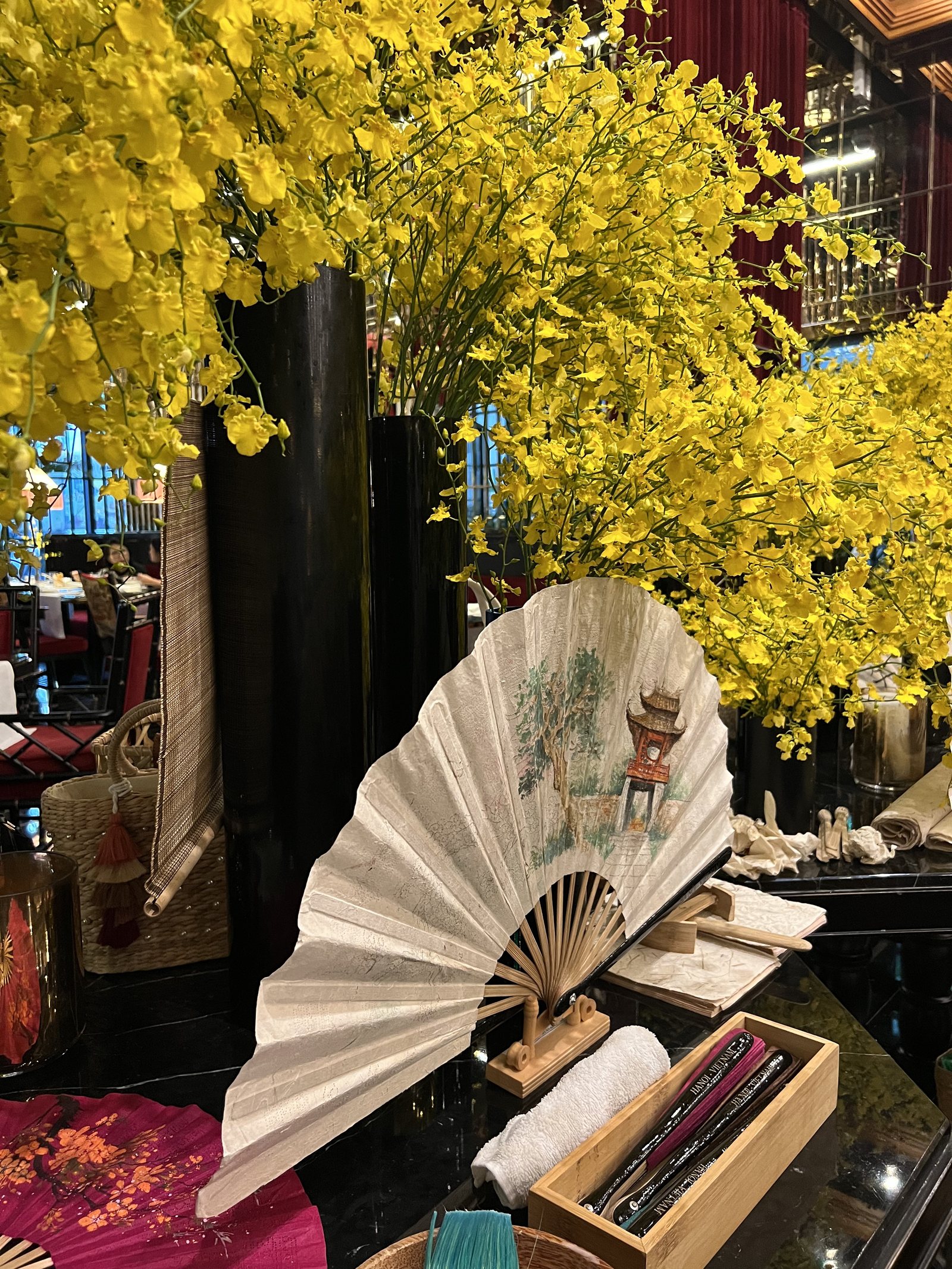 A painted paper fan open on a stand beside a tall vase of yellow blossom