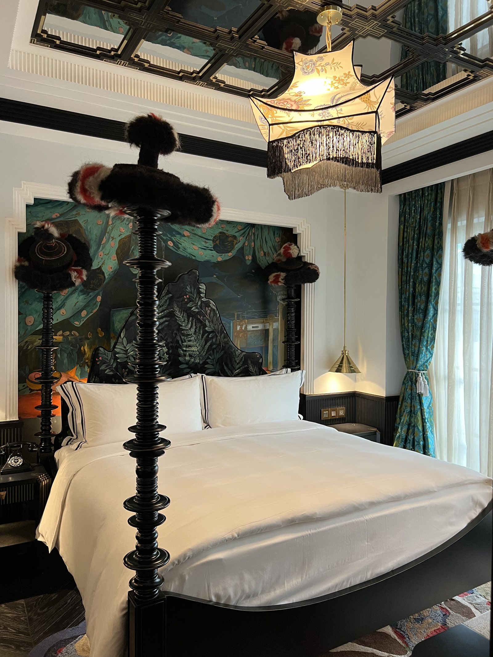 A black four-poster bed with dark feathered finials at each corner, in front of a painted landscape
