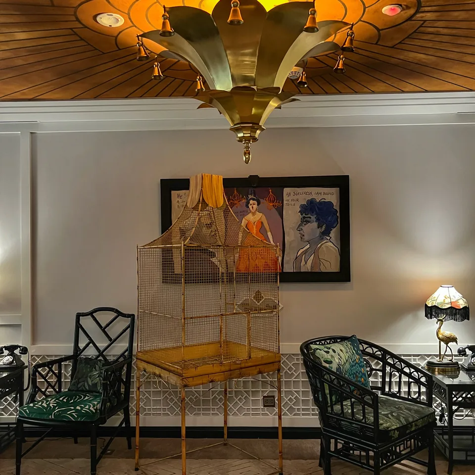 A gilded birdcage on a stand between two painted chairs, under a fan-shaped ceiling light