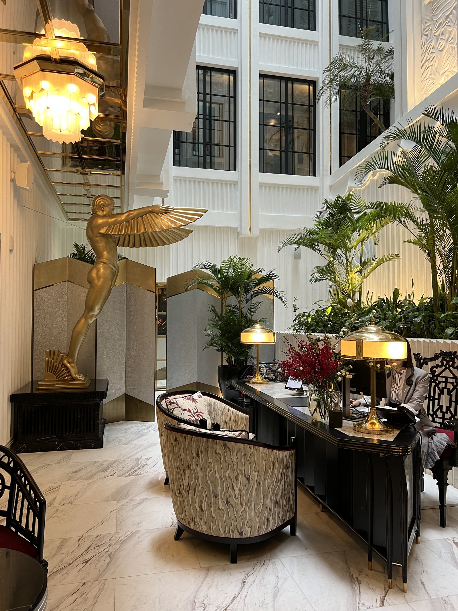 A gilded winged figure on a plinth beside palms, in front of a tall white atrium window
