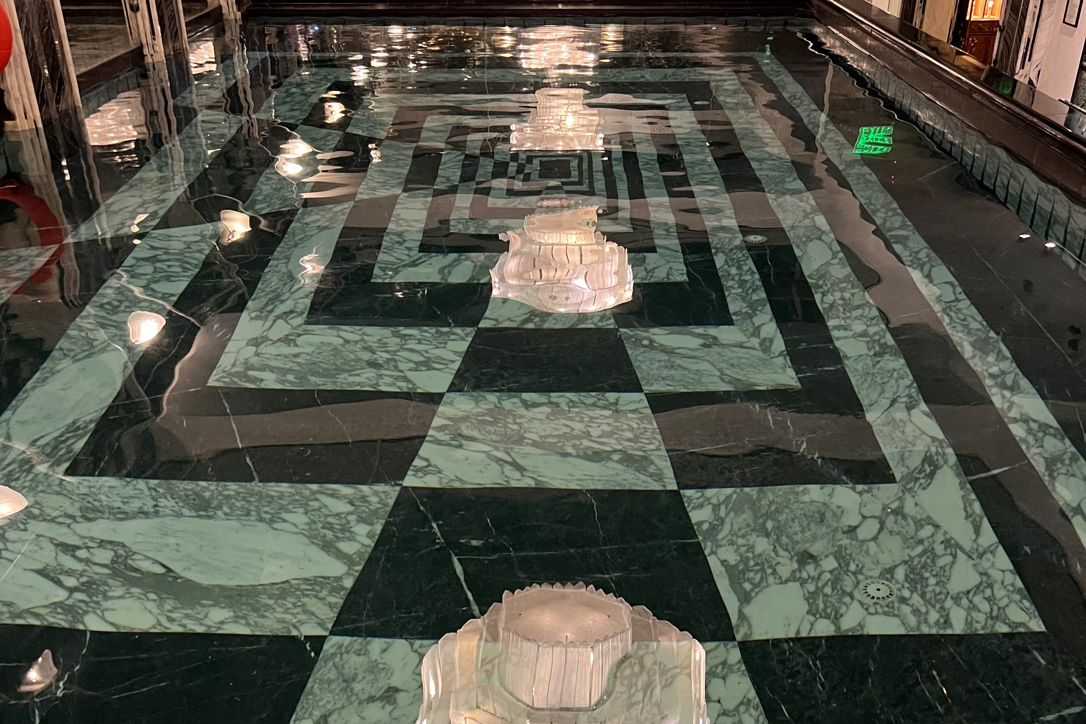 A long indoor swimming pool with green and white marble around it, under a line of lit glass domes and a mirrored ceiling