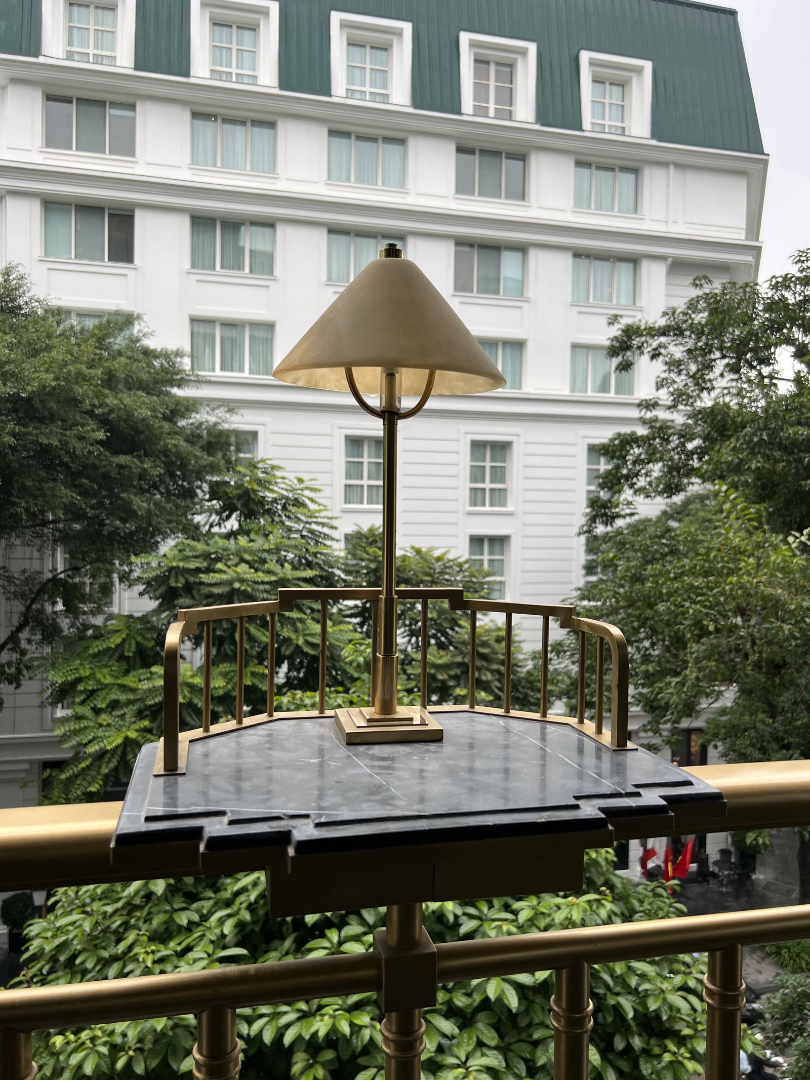 A small balcony with a brass rail and a lamp on a marble table, looking across to a white building with a green roof