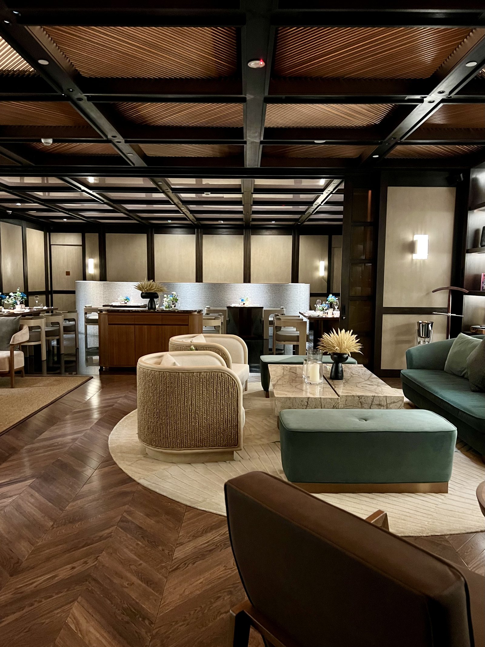 A lounge in pale wood and green velvet under a timber coffered ceiling, with a bar at the far end