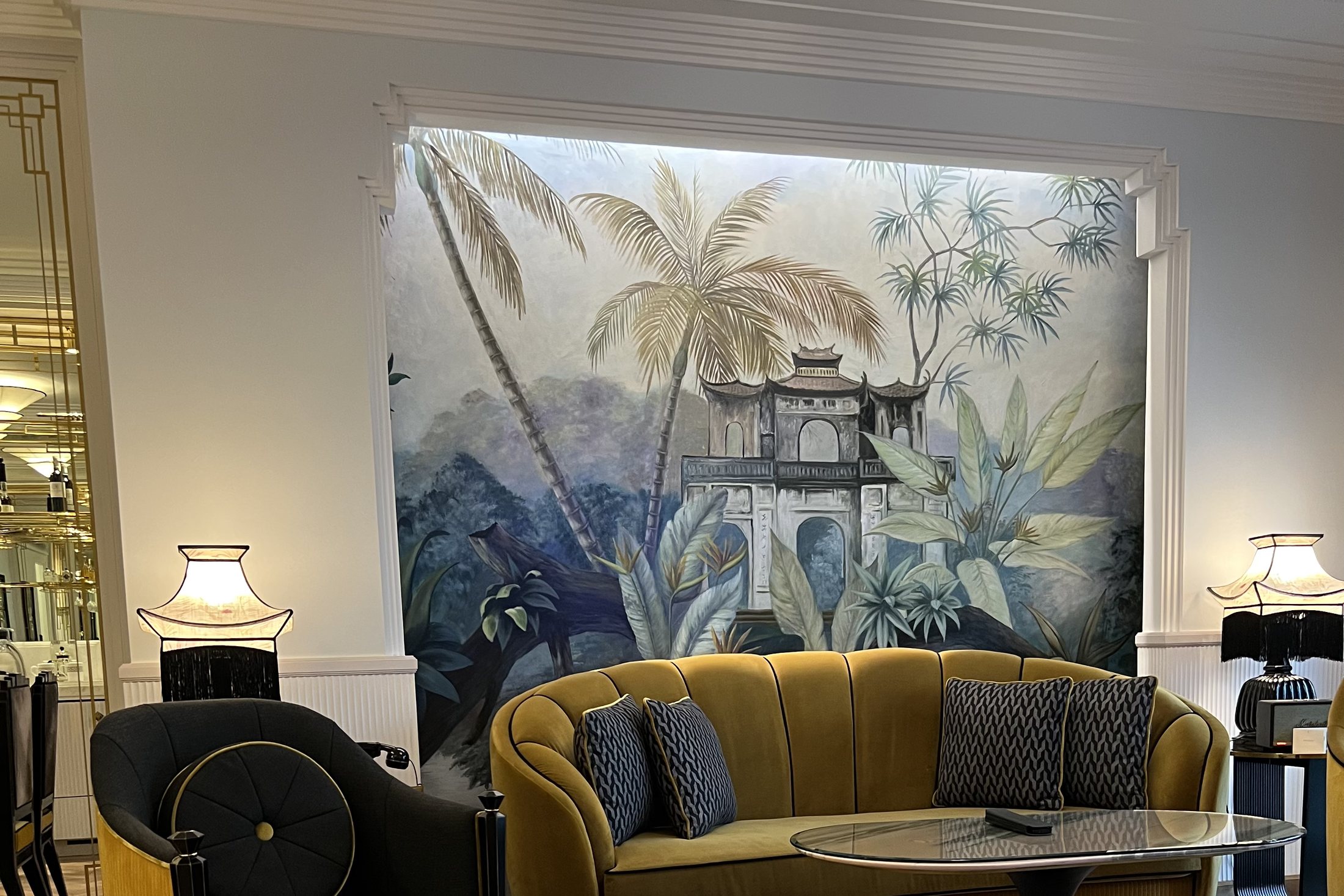 A mustard velvet sofa and a dark chaise longue in front of a painted mural of a temple among palms, on a herringbone floor