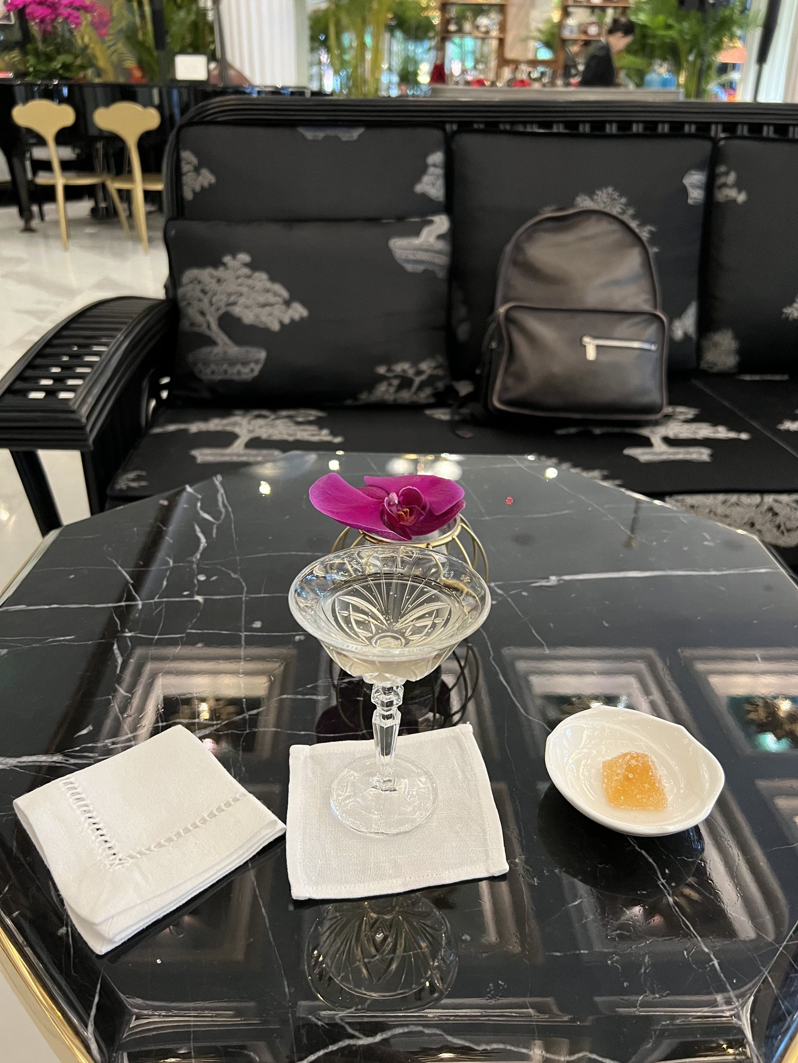 A black marble table set with a coupe glass, a napkin and a small dish, with a backpack on the banquette