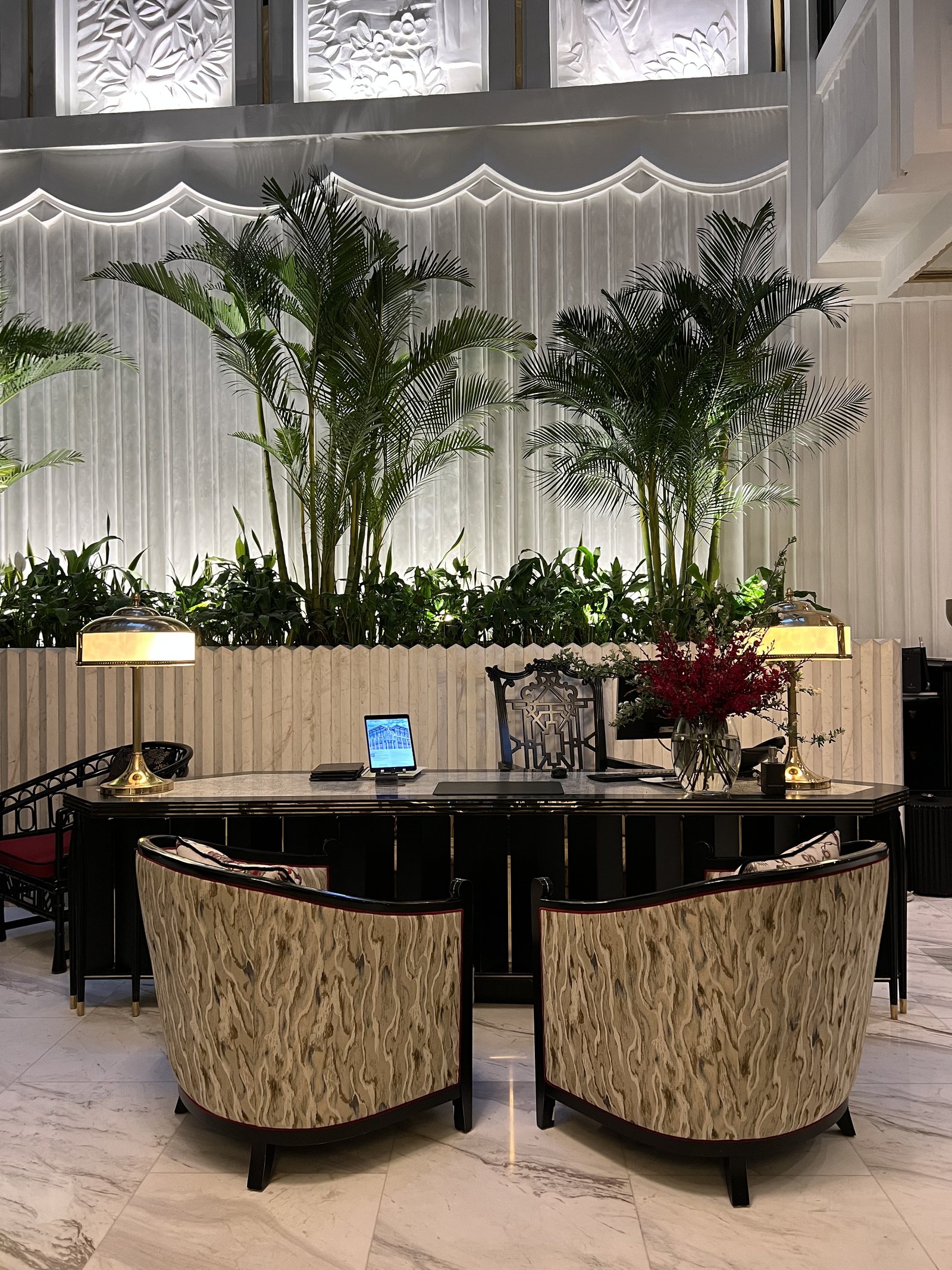 A dark console table with two low chairs in front of it, under lit palms and a table lamp