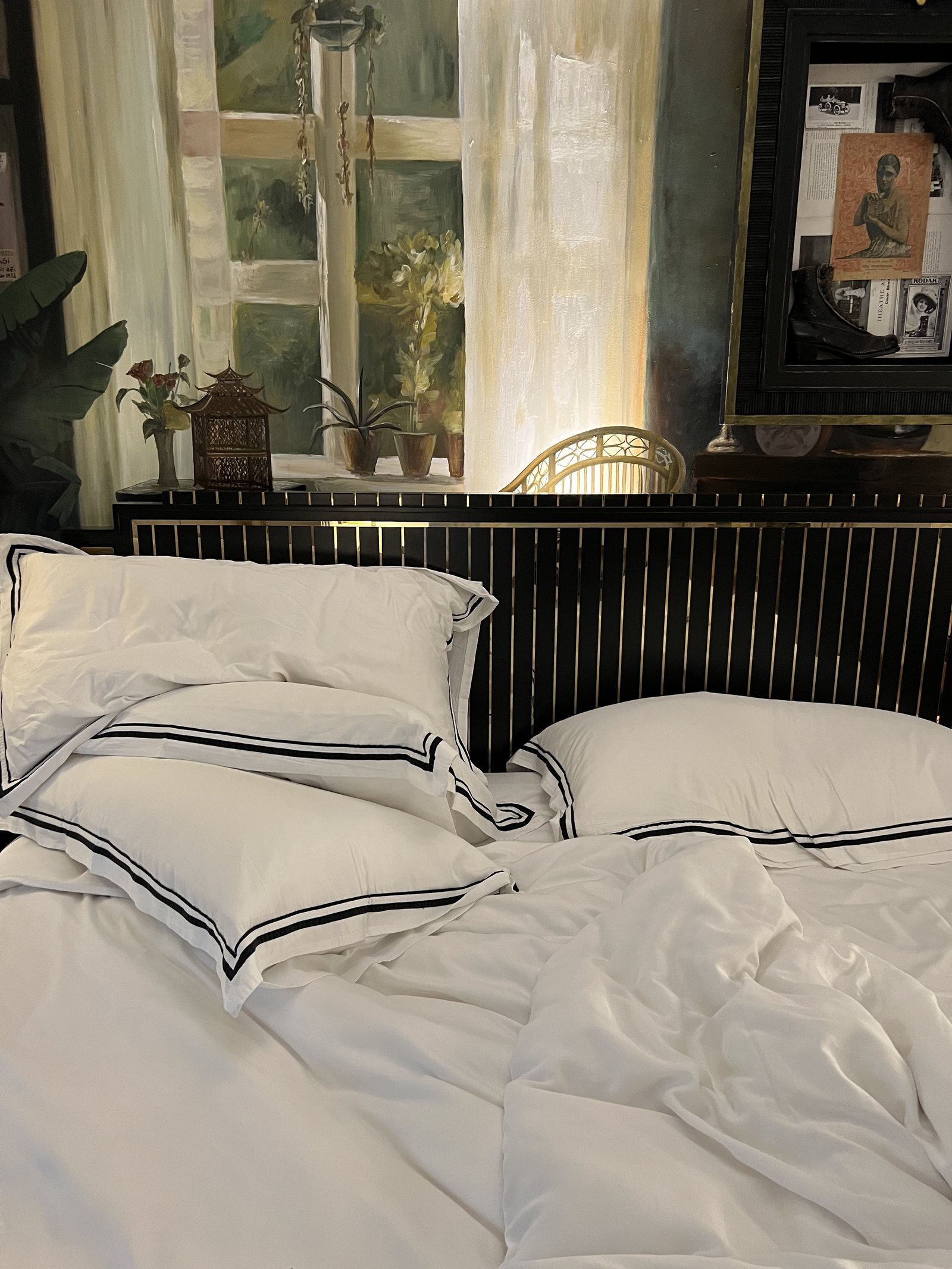 An unmade bed with white linen and black-edged pillows in the morning light, with the painted wall behind