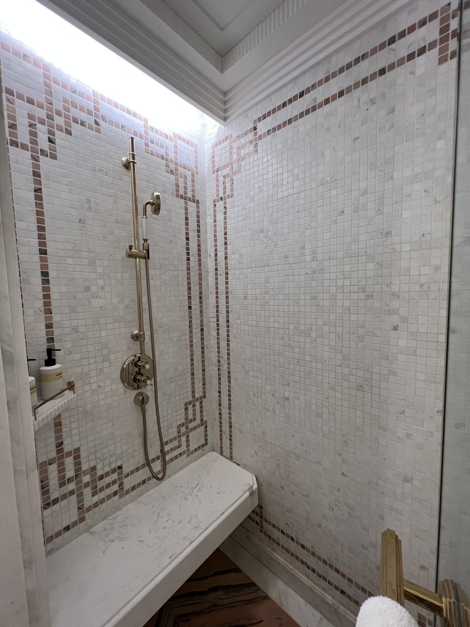 A walk-in shower lined in small white mosaic tiles with a patterned border and a brass rain head
