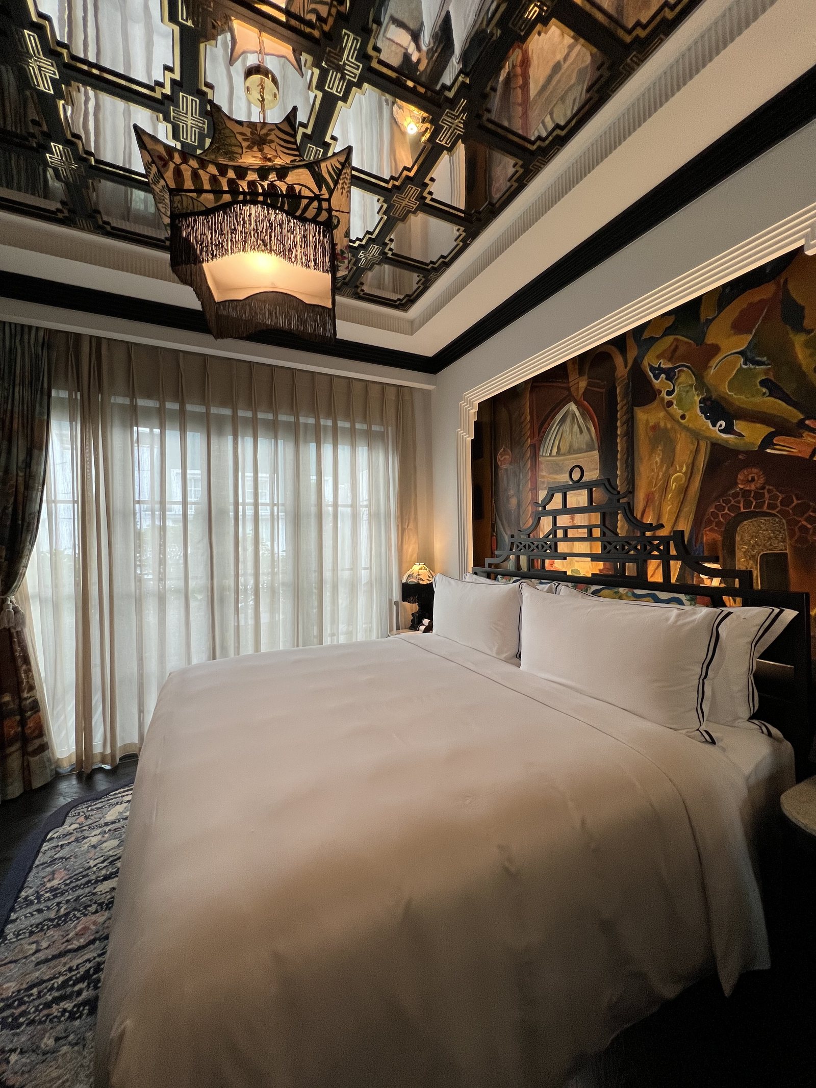 A bed under a mirrored and painted ceiling, with a large painted panel of figures behind the headboard