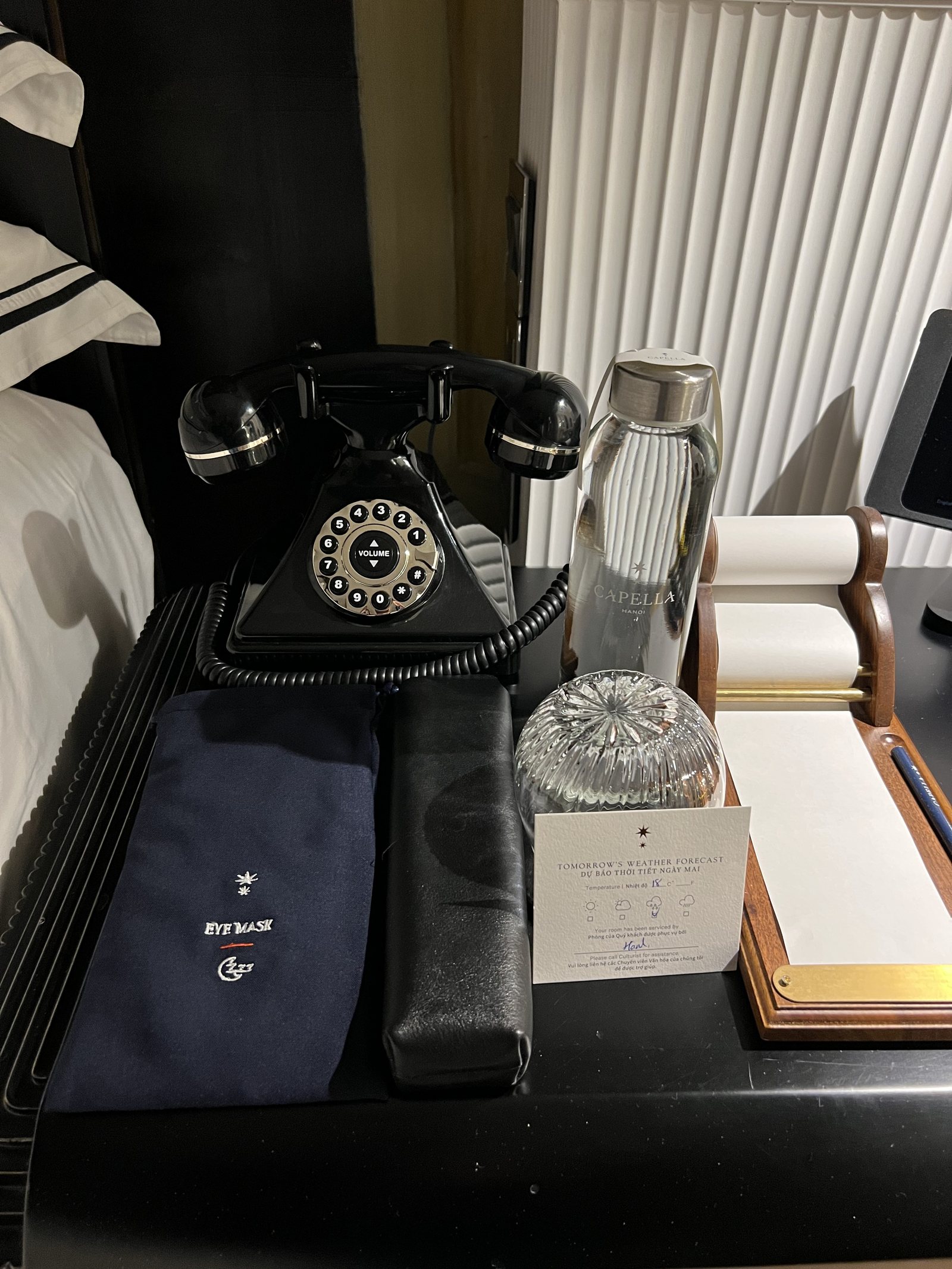 A black rotary telephone, a cut-glass water carafe, an eye mask and a leather roll on a bedside table