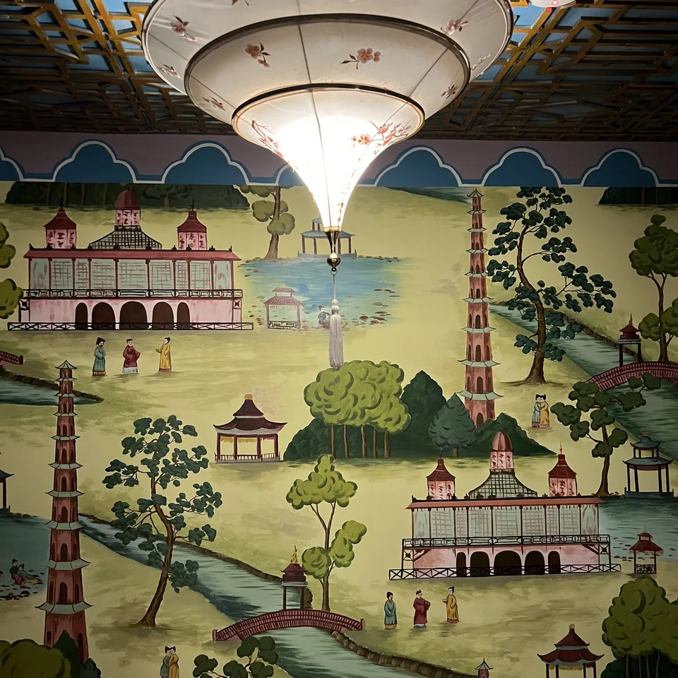 A ceiling and wall painted with pagodas, bridges and figures in a landscape, under a pleated lamp