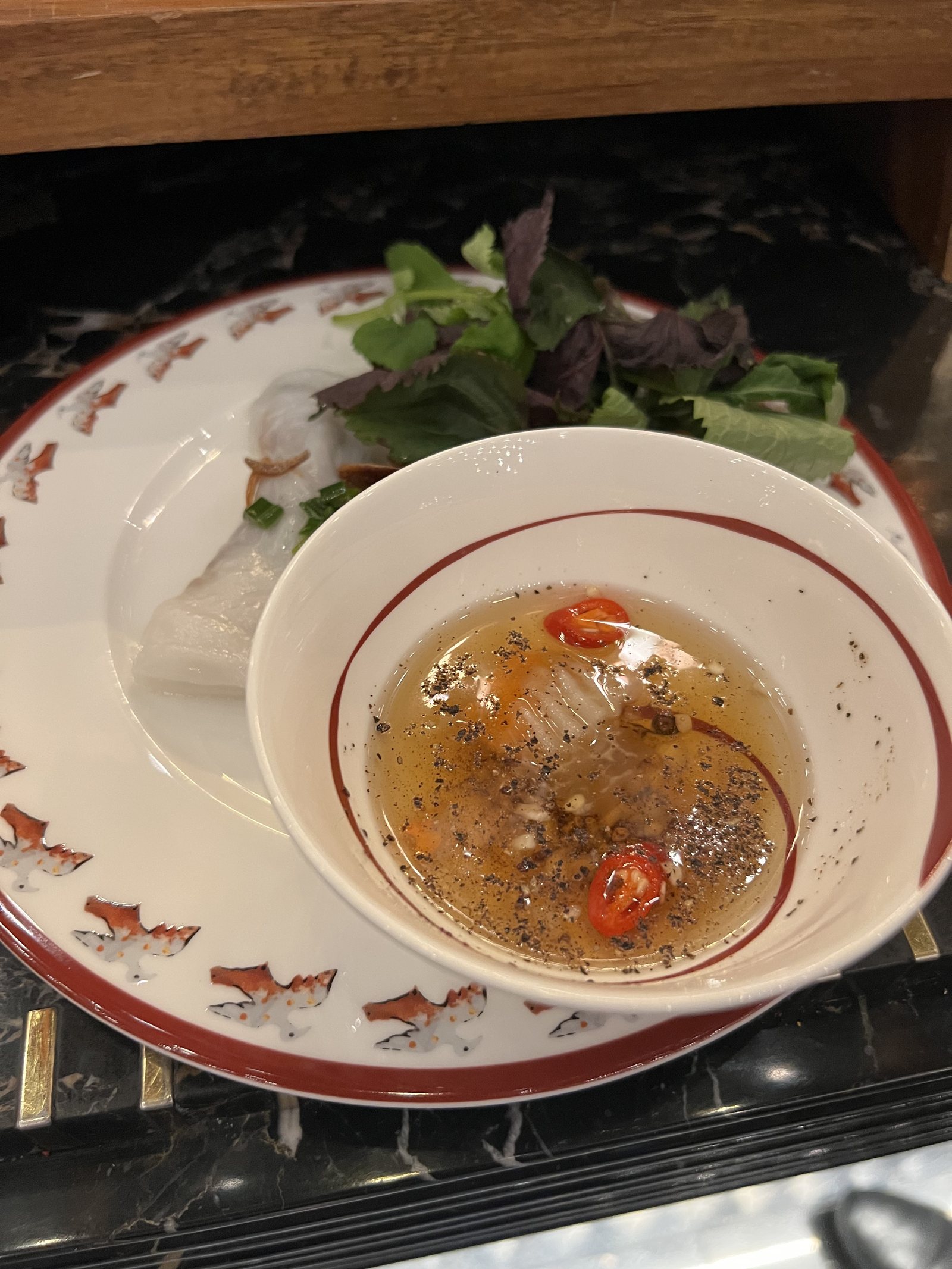 A bowl of clear broth with chilli and herbs on a patterned plate, with a side dish of leaves