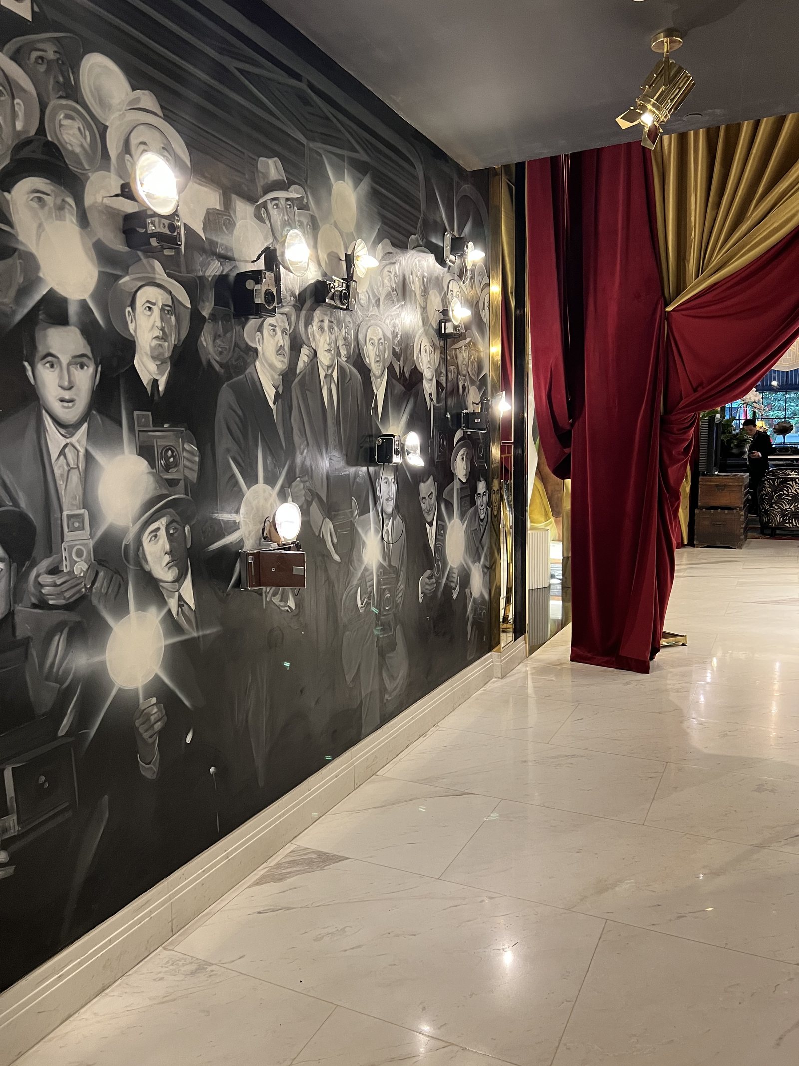 A corridor with a painted crowd of photographers firing flashbulbs, beside a red velvet curtain