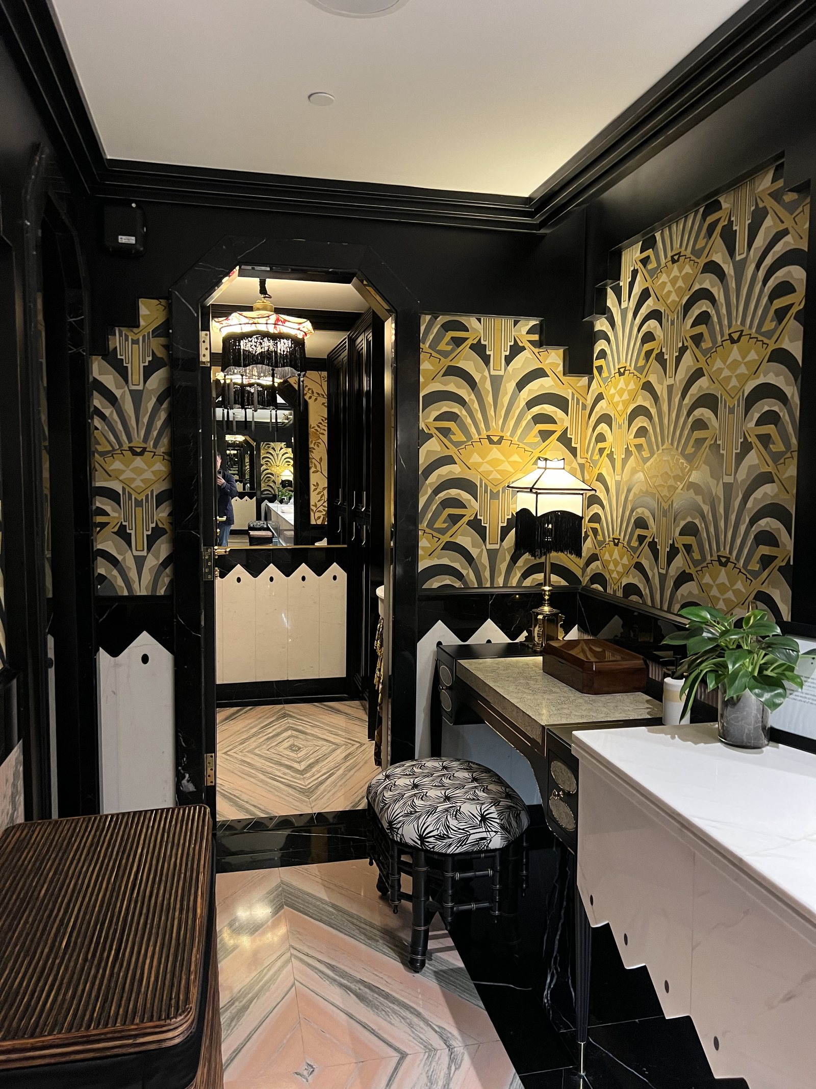 A corridor papered in a gold fan pattern, with a console table, a lamp and a stool on an inlaid floor