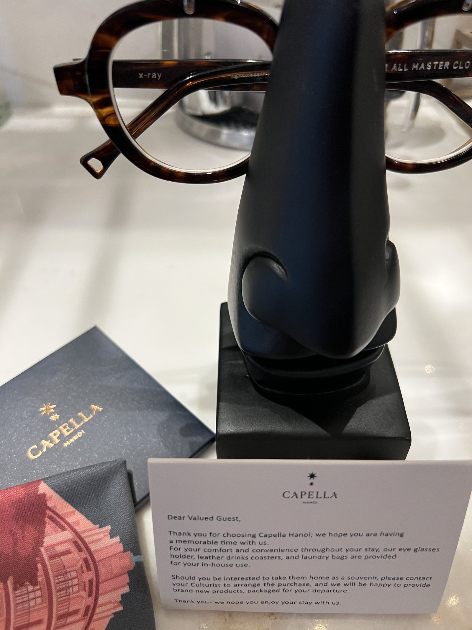 A pair of tortoiseshell spectacles on a black stand beside a Capella box and a printed card