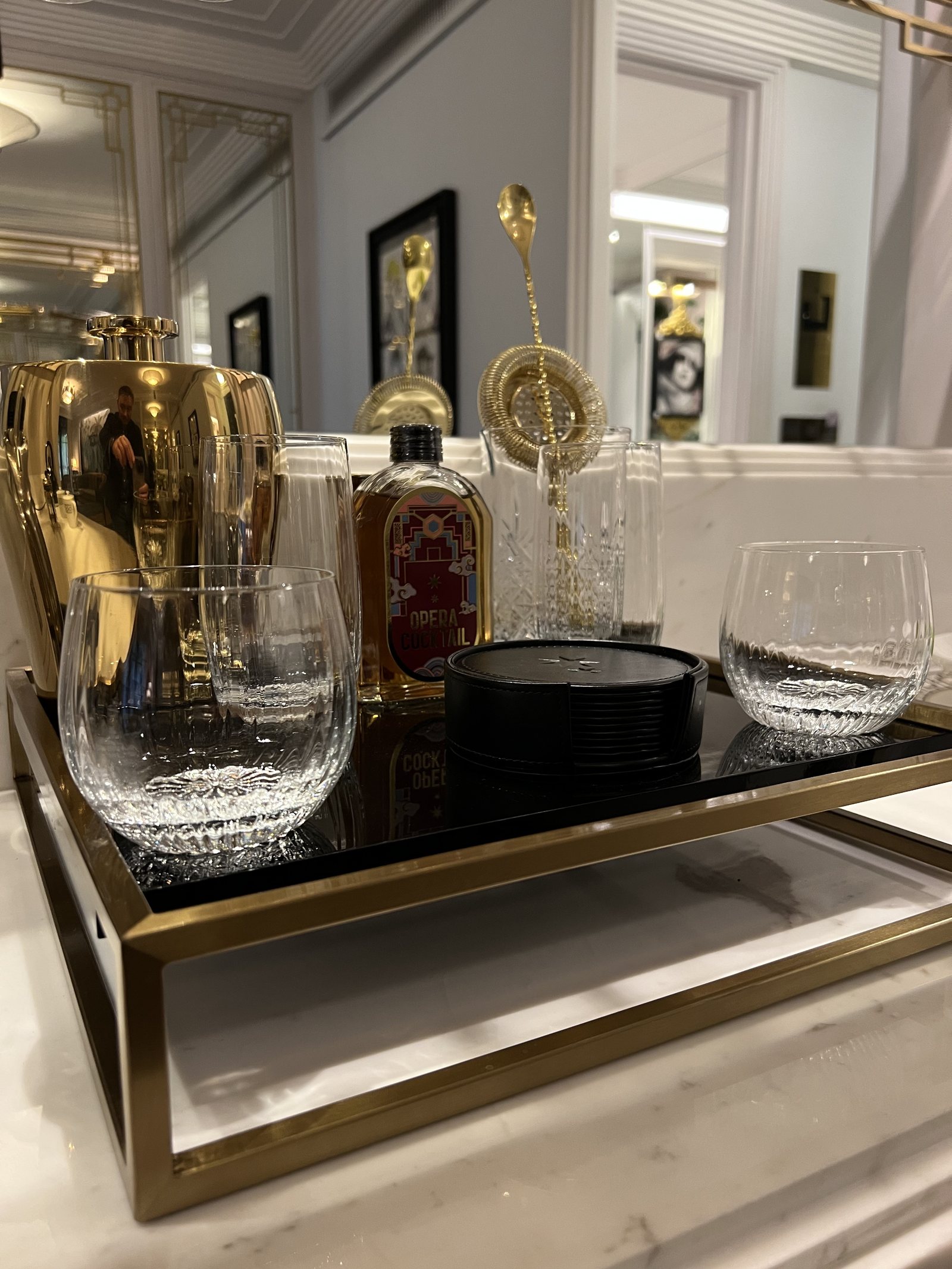 A brass tray holding a cocktail shaker, a bottle of bitters, a strainer and two cut glasses on a marble top