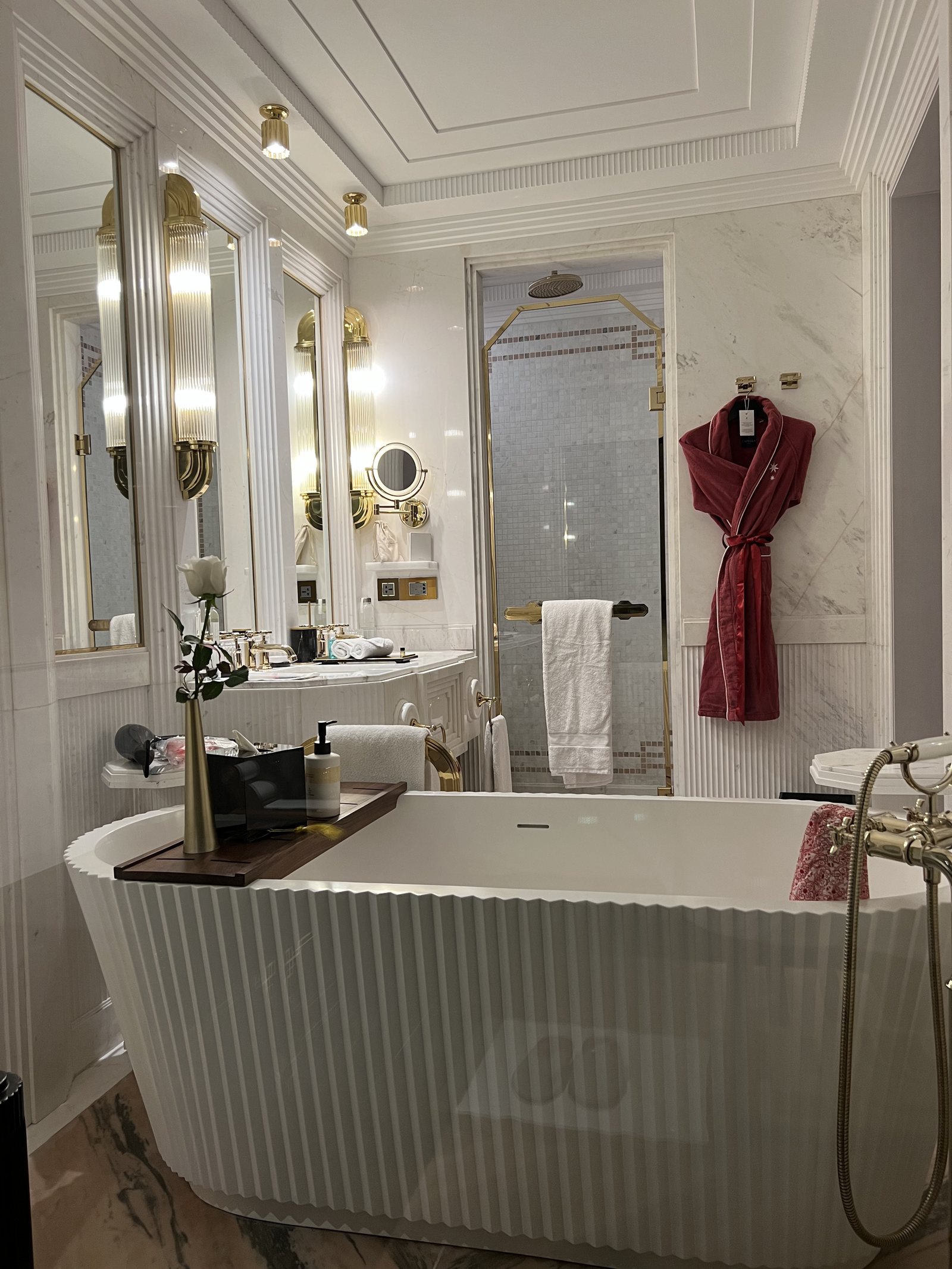 A ribbed freestanding bath in a white marble bathroom, with two lit mirrors and a deep red robe hanging by the shower