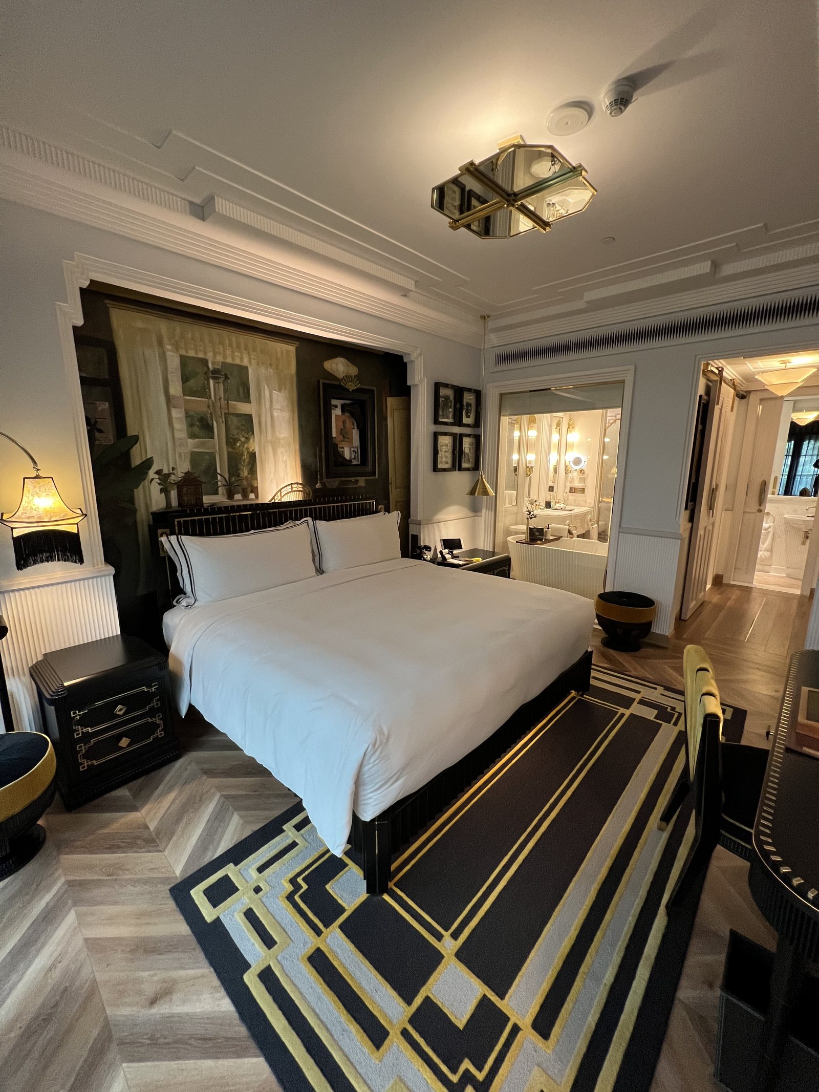 A wide bed with white linen under a tall mirrored headboard, on a black and gold patterned rug