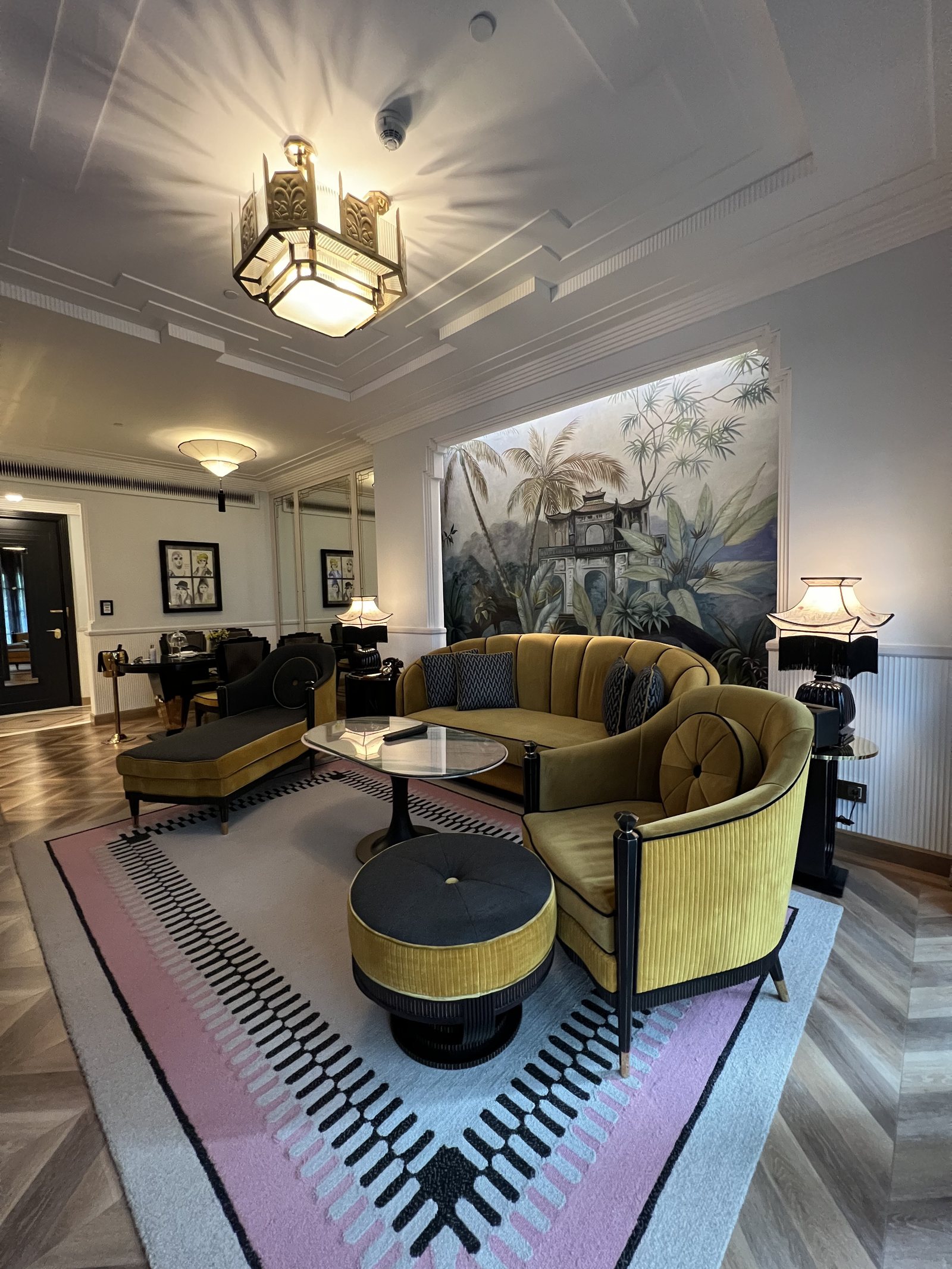 A curved mustard velvet sofa and two stools on a pink and black patterned rug, under a painted panel and a lantern
