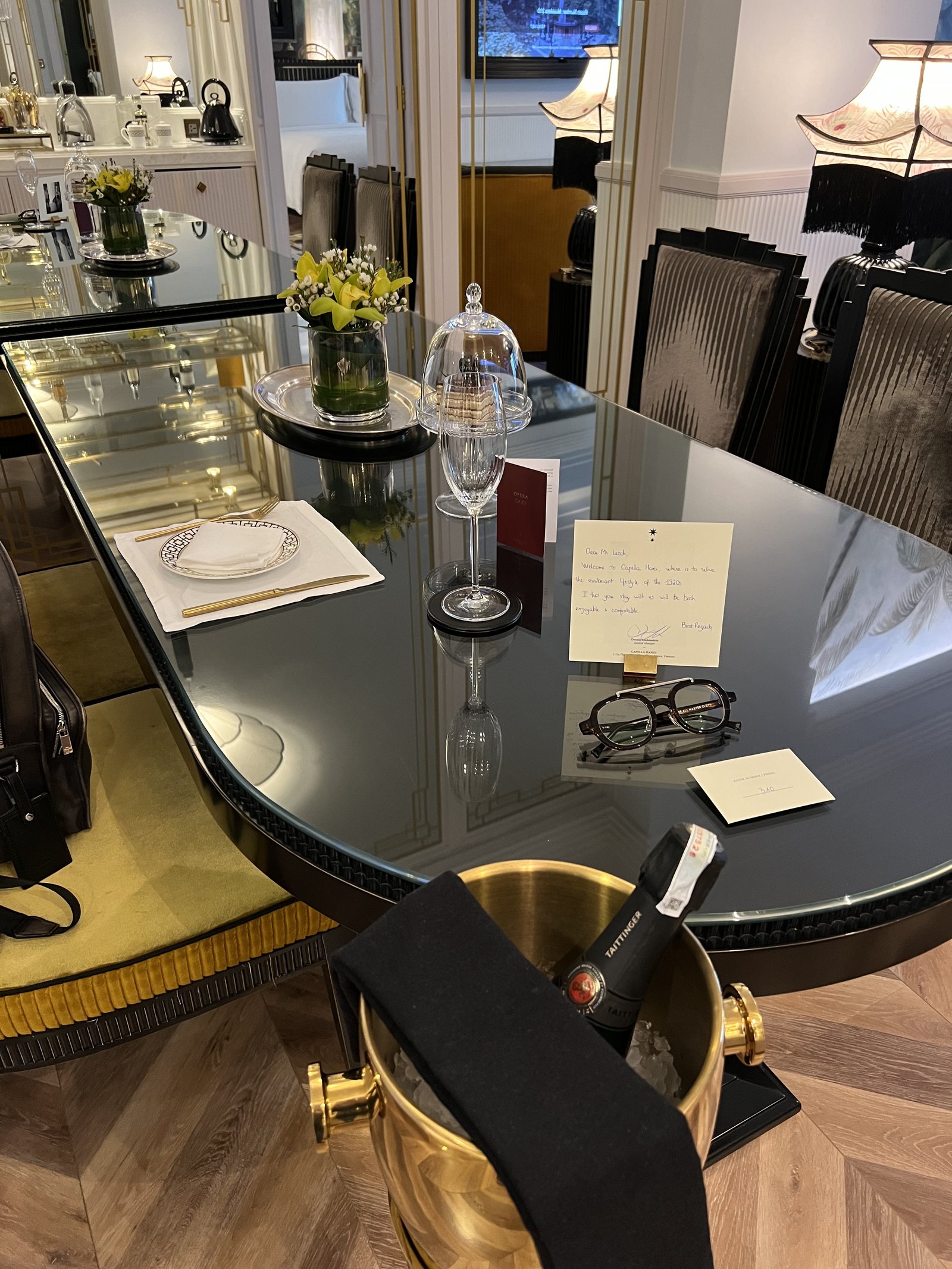 A black oval table laid with a napkin, a coupe glass, a glass cloche, a handwritten card and a bottle in a brass ice bucket