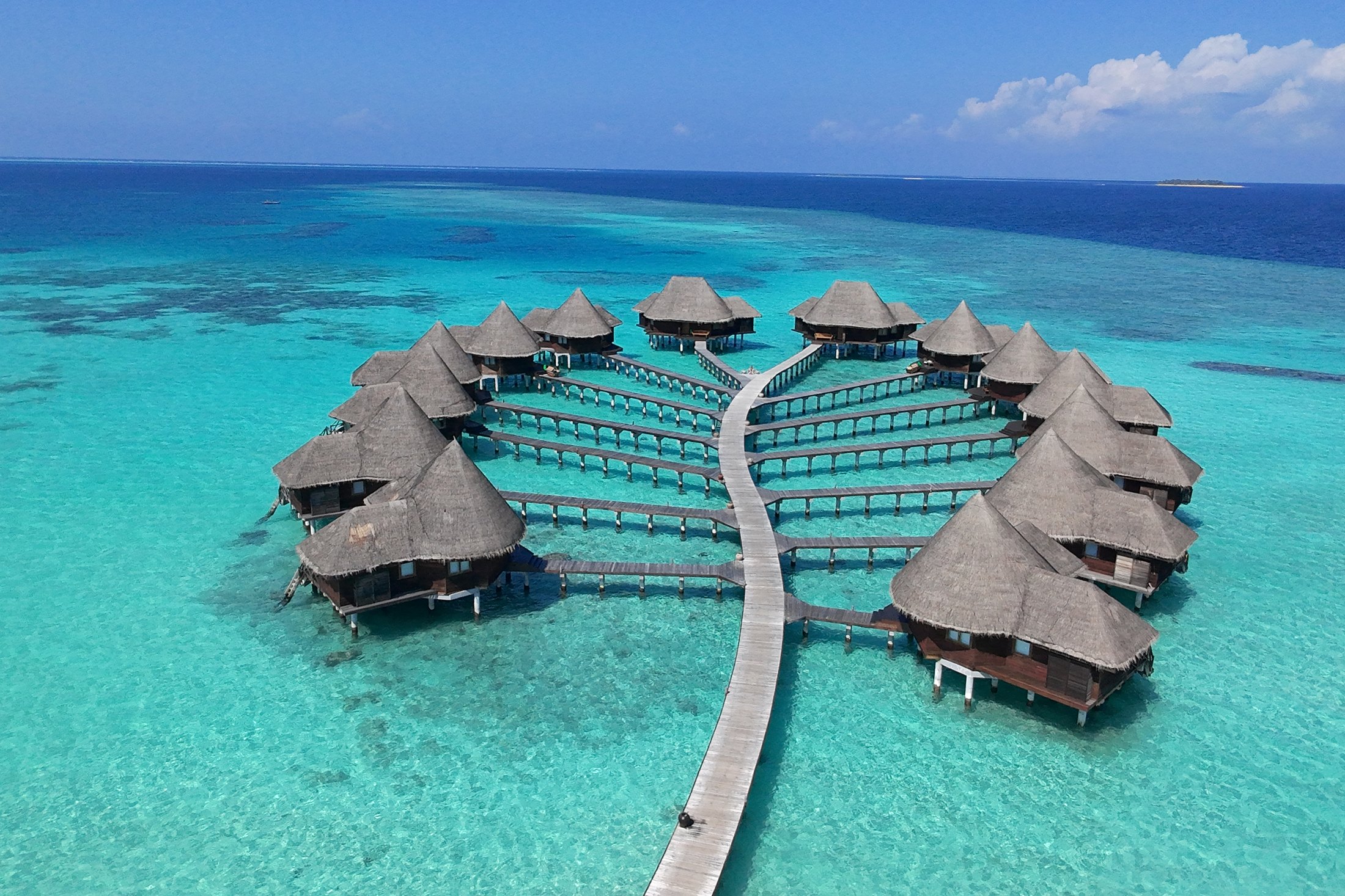 An aerial view of the overwater villas at Coco Palm Dhuni Kolhu, thatched roofs on stilts arranged in a fan around a central walkway over turquoise water, with the reef showing dark beyond