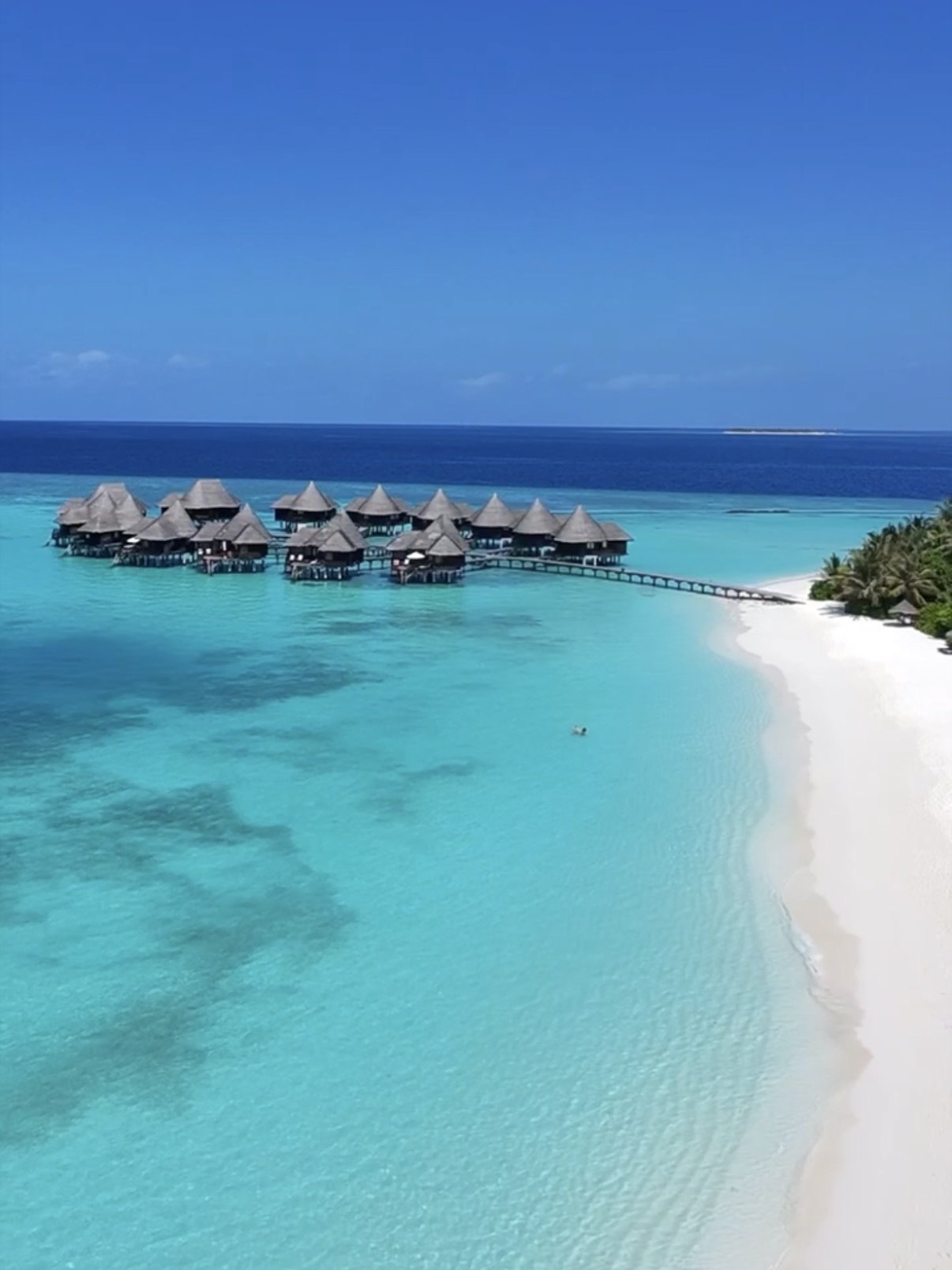 An aerial view at Coco Palm Dhuni Kolhu of the thatched overwater villas clustered over pale turquoise water, the timber walkway running back from them to a wide white beach curving away under the palms