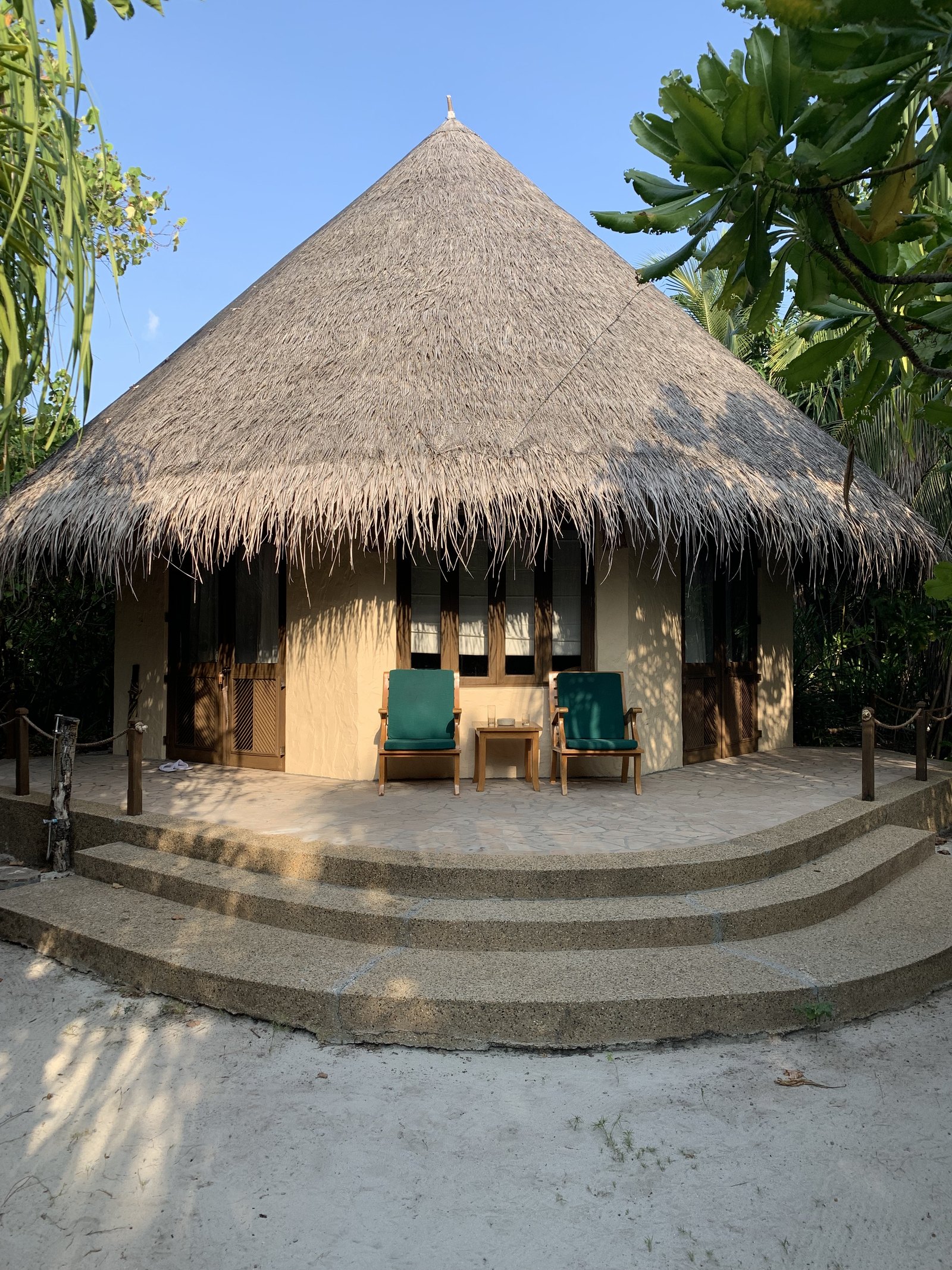 The front of a beach villa at Coco Palm Dhuni Kolhu, a steep thatched roof over a deep terrace with two green chairs and curved sand steps