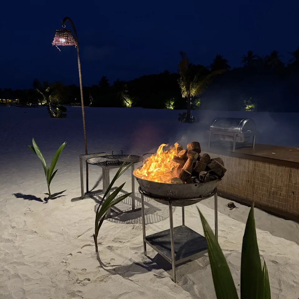 A fire burning in a metal bowl on the sand at dusk, a lantern on a pole behind it
