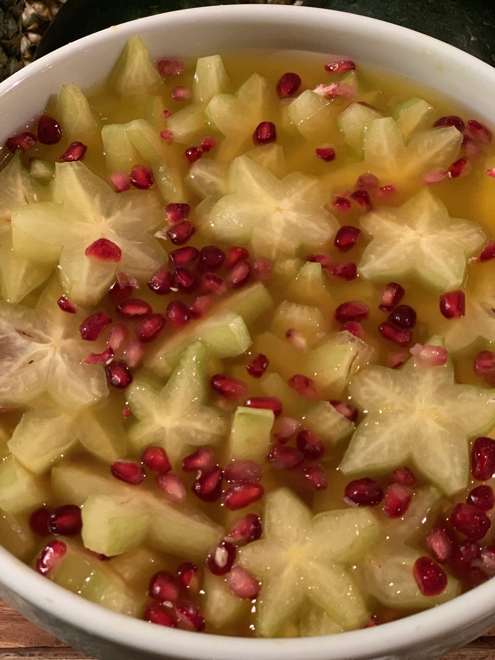A white bowl of fruit salad with sliced star fruit and pomegranate seeds in syrup