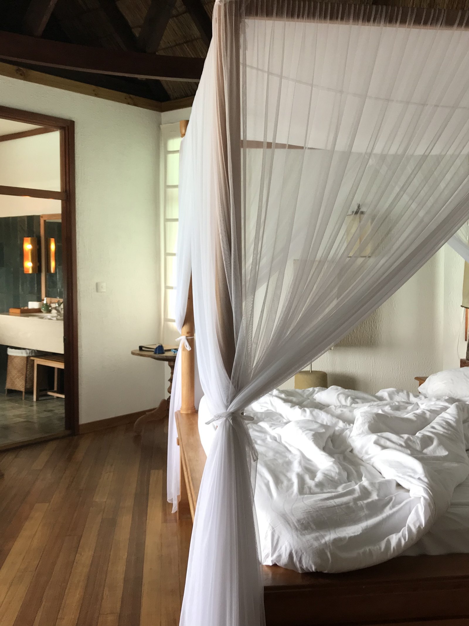 A lagoon villa at Coco Palm Dhuni Kolhu in the morning, the bed unmade under its white net, a standing mirror with a towel over it and the open door of the green tiled bathroom beyond