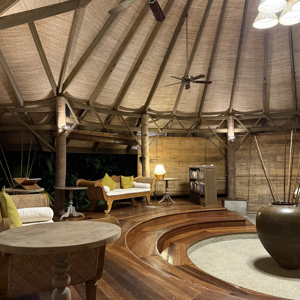 The inside of the thatched arrival pavilion at night, timber floor, a sunken circle of sand and long sofas