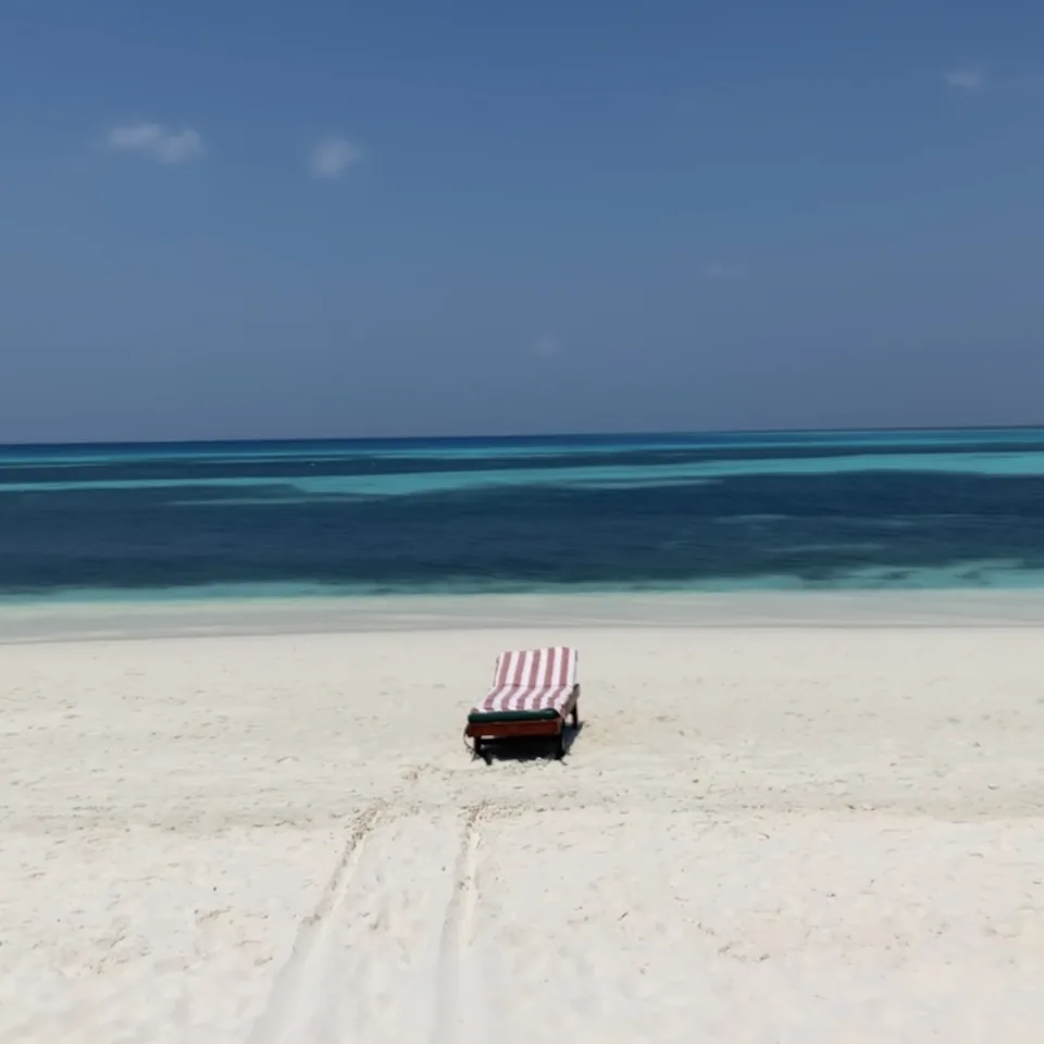 A single striped lounger alone on a wide empty sandbank, the lagoon beyond it